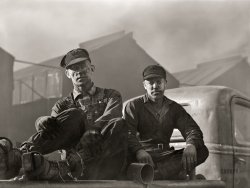 November 1942. "Sunray, Texas. Workers at a carbon black plant." Medium format acetate negative by John Vachon for the Office of War Information. View full size.
The Eyes Have It... and they have Zombie Apocalypse written on them.
Fascinating photoThis is one of those shots that takes the breath away at first glance and, upon further inspection, stuns the viewer silent. The man on the left is riveting for his light eyes with their vaguely troubled and even tiredly challenging expression -- a look which says "Just take the picture if you must." The ropey string tied around his pants legs at the ankles -- is that to keep the dust from getting into his shoes, or did it protect his legs? Whatever it was for, it makes me sad. At any rate, the way his gloved hands are grabbing his shins says "I want you to see this." 
His companion's expression is similarly grave and somehow anxious, with the furrowed brow from the squint and again, light eyes that glow against the carbon that coats his face. He has poetic hands that don't seem to suit the laborer profile. There's a heavy sense of resignation in both of these men but I hope that, despite being weary from hard work and maybe a certain kind of despair at which I can only guess, they were able to get cleaned up and have a good meal and be with loved ones and rest well at the end of each workday. 
Shorpycame to mind.
Getting nothing but staticWould those thick pads strapped to his boots be to prevent static buildup/discharge in an environment heavy with carbon dust?
Unusual footwearWhat is the man on the left wearing over his boots?  I'm not familiar with the process of making carbon black other than it's obviously messy. What a hellish looking place.
Boot attachmentsWhat is the purpose of the attachments to the boots worn by the man on the left?
BlacknessHaving grown up in the same county, I remember the black cloud emanating from the facility when I was a child. Everything was black for hundreds of yards surrounding the plant, including the cattle. Even though the plant is not polluting, it is still in operation and employees are paid overtime every day to take a shower. It is not as bad as in 1942, but the blackness still permeates.
Shoe Blocks?The blocks attached to the shoes -- is that to protect from heat?  I find it interesting that the workers took great care to protect legs and arms with tight fit but no protection from breathing the dust was apparent from the very dirty faces and nostrils. 
(The Gallery, Factories, John Vachon)