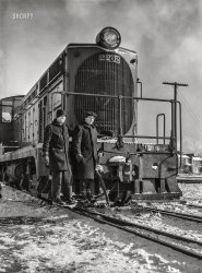 March 1943. "Chicago, Illinois. Switchmen riding one of the Atchison, Topeka and Santa Fe diesel switch engines." Photo by Jack Delano for the Office of War Information. View full size.
Tough GuysYou do not want to be a hobo riding the rails and run into these two!
No expense sparedNote that the AT&amp;SF went with fancy raised metal numbers on the engine's number plate when Baldwin built this engine for them in 1939.  Simply painting the numbers would have been a lot cheaper.
ColdWool pants and coats, heavy mitts, hats over the ears – it’s definitely cold here.  Plus I like the twin ciggies.
Baldwin VO-1000According to my ATSF all-time diesel guide, #2202 was a Baldwin VO-1000 delivered in 1939. Prior to seeing this photo, I didn't know that the Santa Fe received VO-1000 locos that had an oval radiator grille instead of the later rectangular one.
Baldwin 62303The build plate is next to Lefty's right glove. No. 62303 was Baldwin's third VO-1000 diesel, completed 12/12/1939

Link &amp; Pin Days RemnantIt's surprising to see that the knuckle of the coupler has the slot across its face that would have allowed it to couple with the link &amp; pin coupler system outlawed around 1900. The early Janey couplers came with the slot for the transition period between the systems. That slot weakened the knuckle and they failed to the point that some suggested a return to the link &amp; pin. Fortunately, the railroads were making more money hauling longer trains with the new coupler and the number of brakemen losing fingers and hands started to recede from the tens per day that had been the norm.        
Love those coats... and if you've never experienced the warmth of winter weight wool pants on a cold Northern day, you're missing out.  Our ancestors knew how to handle the weather in a way we too often do not.
Yeah, it was coldThere's no fashion involved for those two - March of '43 started out colder than normal and those two knew how to keep warm. I bet they had some good wool underneath as well.
Baldwin DieselsBaldwin was a large successful manufacturer of steam locomotives that struggled to make the transition to diesel power and eventually exited the market in the mid 1950's.   
This early offering illustrates some of Baldwin's thinking including an indestructible cast steel frame, the cast numerals for the locomotive number, and the electrical receptacles on the top corner of the front hood that could be used to plug in marker lights attached to the side brackets.   Baldwin would soon economize this design with a flat grille for the radiators and a painted road number.  Here is sister 2252 near the end of her career in 1966 with different trucks and a rakish multi-stack exhaust.   https://www.railpictures.net/photo/392182/ 
Riding the footboardsOnce a very common practice, it was obviously quite dangerous - one slip when riding on a moving locomotive would put you directly under the wheels, which as you can imagine led to a horrific result.  The practice was eventually outlawed in the 1970s and all footboards on existing locomotives removed by around 1980.
(The Gallery, Chicago, Jack Delano, Railroads)
