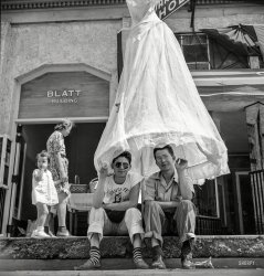 July 1940. "Street scene at the fiesta in Santa Fe, New Mexico." Our third look at the festive goings-on here, and we'll let someone else explain the skirt-brella. Photo by Russell Lee for the Farm Security Administration. View full size.
Wicked TeeWhen did screen printed t-shirts become a thing? I googled about and the technology existed, but Wikipedia glossed over the early days of screen printing and went right to the 60s. 
Plus, does anyone know what the shirt is talking about? The Santa Fe Devils? Demons? Beelzebubs?
Never mind the skirtI want to know about those socks!
Blatt Building, Santa FeIt took a bit of digging, but I found where this was taken (roughly). It's now called the Silver Building, and still retains the funky trim visible in the photo.
View Larger Map
Do not repost this photo!If it goes viral then every hipster in America will be wearing those sunglasses.
Never Mind the SocksThat's an interesting variation on aviator's sunglasses. Besides the dress, which may be hanging there advertising a sale or something of that nature, there's a general oddness to this picture that I can't put my finger on.
Decent ChapsNeither is looking up.
Holey HuarachesNever mind the sunglasses -- check out those Wicker Slippers! I wonder if they come pre-tattered?
&quot;Ankle Beaters&quot;When I was in school if you rolled your pants up too high you risked verbal humiliation.
Possible &quot;skirt-brella&quot; explanationI think one of the previous commentors is on the right track with " the dress, which may be hanging there advertising a sale or something of that nature". It may be advertising the "White House in the ground floor of the Catron Block. ... Santa Fe’s first ready-to-wear, woman’s fashions. The Blatts eventually purchased the Catron Block, renaming it the Blatt Building."
Petticoat JunctionI got it!
Be True to Your SchoolSanta Fe High School is still at 2100 Yucca Street, and its team mascot is still the Demons. He is probably a student there and that is an athletic or school spirit shirt.
Ladies wearMy source is not entirely clear, but there was a women's wear store called the White House at that location through at least the '30s.  The sign mostly obscured by the dress at the top of the image is more consistent with Guarantee Shoes, next door.
How many names does it have?Using Sagitta's link to the Google street view, you can zoom up to the crest of the building and see that it still says Catron Block, named for the notorious land grabbing, Republican Ring lawyer Thomas Catron, who died in 1921.  The law firm still does business in their Santa Fe office, and there is a county named after him in NM.  The Catron Block sits appropriately opposite the Palace of the Governors on the ancient Santa Fe Plaza. 
HuarachesThose are called huaraches, a type of pre Colombian Mexican sandal. They were HUGELY popular among young people when my mom was in college, (1937 - 41).
(The Gallery, Russell Lee)