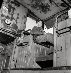 March 1943. "Walter V. Dew, rear brakeman, on the Atchison, Topeka &amp; Santa Fe between Chicago and Chillicothe, Illinois, watching the train from the cupola." Photo by Jack Delano for the Office of War Information. View full size.
Essential RR EquipmentCigar boxes.
Old time ingenuityClever use of cigar boxes.  Today the brakeman would complain to management and the union.  After six pages of paperwork, someone would be designated to hang a couple of metal boxes.
No maintenanceThe railroad didn't spend too much on frills (paint, shelving, etc,), did it?
&quot;You lookin&#039;at me?&quot;Mr. Dew looks like he does not care to have his picture taken!
You never knowwhen half a shoelace will come in handy.
A quizzical smile on his face, as if to say you're down there and I'm up here, in charge.
My FatherWorked for the railroad in the 1930s. He's not with us now but just recently I found out that a pitchfork of his that I use to spread mulch is actually a coal fork, he probably acquired it while working the railroad. It has R&amp;F stamped / burned into the handle. Looking at this picture I noticed those square nuts holding parts of the cabin the brakeman is sitting in together. I have tins of these my father had in his hardware stashes and had never seen them used before. They're the same, flat on one side and sort of curved on top. He worked outdoors on the mechanicals of the train. He used to say that a lot of the guys that worked the boiler shops were gone by the 1950's, they handled a lot of asbestos I guess. He used to think this was the reason.
No frillsDuring the war the maintenance was to keep the trains running and the materials flowing. Taking a caboose or any other needed equipment out of service for cosmetic work just wasn't done 
About that cigar boxBack when my father-in-law passed on, I was given the enjoyable task of cleaning out his basement and I found a number of old cigar boxes from HIS father. Back in the late 1930s and 1940s, these were made from a lightweight wood, unlike the heavy cardboard of today, and from what I saw in that basement, they were for a number of aftermarket uses. This photo just shows another one.
Slowing down (probably)One of Walter's jobs was to keep an eye on that gauge, which shows the air pressure in the brake line.  Normally (brakes off) it'd be at something like 80 or 90 pounds.  When the engineer applies the brakes, the reading would go down; here it's 74 pounds, which probably means at least a little brake application.
If the gauge dropped really low really fast, it probably meant that a brake hose was open, either because the hose burst, or because the train had broken in two.  If it dropped somewhat low and stayed there, Walter knew to hang on, because the engineer was stopping hard for some reason.
Today, this monitoring function is done by a box clamped to the coupler of the last car, called a Flashing Rear End Device or FRED.  The brake hose on the last car is plugged into the box, and it transmits the current pressure by radio to a display in the cab.  (Some engineers call the cab display box "Mary".)  As the name implies, FRED also contains the flashing red light that you see on the end of a train at night; it comes on automatically when it gets dark outside.
Chicago and North Western LineIn the King Edward "Invincible" cigar box we may find the maps the brakerman used. The "Chicago and North Western" map we find on top, as we can see the "ball and bar" logo.
According to the Wikipedia entry on CNW, in 1943 the text must have been "Chicago &amp; North Western Line" (1902-1944).
I suppose it was an "Employee Owned" map. here you may find one of 1977. I couldn't find one of around 1943. 

Too Gone Too LongAs a rail fan, Walter is on my list of screen saver images and each time his gaze comes round I find Randy Travis looking back at me.
What is missing.As a kid in the 1950s I rode in several cabooses.  There were girly pictures on the walls of the cabooses.  None here in this pic.  Perhaps removed for the portrait?
[There are plenty of girly pics in Jack Delano's other caboose shots. - Dave]
(The Gallery, Jack Delano, Railroads)