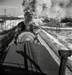 March 1943. "Chillicothe, Illinois. A helper engine is taken on for added power on a grade extending eight miles on the Atchison, Topeka &amp; Santa Fe between Chillicothe, Illinois, and Fort Madison, Iowa." Medium-format negative by Jack Delano for the Office of War Information. View full size.
Coordination?I wonder how they coordinate power levels, or maybe the lead engine goes all out and the trailing one controls the speed.
Must Be a Steel CabooseIn earlier times, a wooden caboose was placed behind the pusher. Leaving it between the loco and rest of the train could end badly for the tail end crew.
Risky BusinessUse of helper engines was a common but dangerous practice prior to automated controls. My father, a long-time railroad special agent/claims agent, listed a couple of helper-caused wrecks as two of the worst he had to deal with.
Helper Engine#3286 was one of 128 locomotives ordered from the Baldwin Locomotive Works, Philadelphia, Pennsylvania. The locomotives were built between 1917 and 1920, and delivered in batches, with 3286 being the next to last delivered.  The Santa Fe was a steady customer of Baldwin-built locomotives over the years.  3286 was a 2-8-2 Mikado Class engine.  All of the 128 locomotives of this class were gone by 1955.
Minor Refinement to LocationGreat photo!  The location is where the Santa Fe (now BNSF) crosses the Rock Island (now IAIS). The train is starting on the climb up the 1% Edelstein Hill.
As a child I spent a lot of time at this exact place watching freights &amp; the Chiefs. (Born too late for stream.)  The best visit was a late night Thanksgiving with a gentle snow &amp; 2 caboose back to back with flickering oil lanterns.  (Which surprised me since I always thought they were electric.)
Considering the year &amp; month,  it is probable my grandfather was clerking at the station &amp; my Dad was working on the bridge in the background over the Illinois River.
Pusher manAfter the helper is coupled, there is a simple application/release air test.In preradio days, we'd hold off on the release until we were ready to go. When the air released, the helper would start shoving and the lead engine would start pulling. You'd be amazed how fast a long freight can get up to speed from a standing start with engines on both ends.
The lead engine aways controls the speed. The helper's job is to push.
(The Gallery, Jack Delano, Railroads)