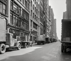 March 1943. "New York. Trucks in the garment district." Medium format nitrate negative by John Vachon for the Office of War Information. View full size.
Now and ThenGarment center is now mostly farther west.
View Larger Map
Governor Clinton Hotel - 1962At the age of 8, I stayed with my family in the Governor Clinton Hotel, and was fascinated looking out the window at the men pushing racks of clothes down the street wreathed in clouds escaping steam.
What is the nameon the trailer on the left? It looks like FREUHAUF or is the last letter an R? Freuhauf is the current name so I expect it is an F
Looking at these pictures I find it interesting how long some brand names have been around.
[Fruehauf Trailer Co. was in business from 1918-1997. -tterrace]
Millinery DistrictWest 36th Street in this area was part of the Millinery District.  A subset of the larger Garment District, the Millinery District ran between Fifth and Sixth avenues and from 35th to 40th streets. During its heyday in the 1920's it housed over 600 hatmakers with a total of 15,000 employees. It already was in decline by 1943, and today only a few specialty hat businesses remain.  The name is still commemorated by the Millinery District Synagogue on Sixth Avenue between 38th and 39th streets, though being on the west side of Sixth the temple is actually just outside the district's boundaries. 
Railway ExpressThe fourth and fifth trucks in line appear to be Railway Express trucks, the UPS or FedEx of the day.  At least they didn't park in the middle of the street like modern delivery services do.
The semi on the leftI just love the appearance of that semi on the left. What make was it?
InternationalThe first truck on the left hauling the Fruehauf Trailer looks like a 1941 - 1943 International.  Most likely it is a 1941, but some civilian truck production continued throughout the war in order to help move goods to market.  The model may be a 5-Ton K-8 which was very popular.  This model " . . . used a Red Diamond 318 engine and a five speed overdrive transmission with single or double reduction axles (K-8, KR-8) or a two speed axle (KS-8)."  The windshield still opened up at the bottom to allow additional ventilation in the cab.
Source: International Trucks by Frederick W. Crimson, p. 170
(The Gallery, Cars, Trucks, Buses, John Vachon, NYC)