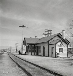March 1943. "Isleta, New Mexico. The Santa Fe depot. Horizontal arms on pole indicate a 'red beard,' that is a message is to be picked up by the train crew." Photo by Jack Delano for the Office of War Information. View full size.
MultitaskingThe indication also required (I think) the engineer to step into the office and sign for the orders.
It was also used in a not-by-the-book way by the station operator to slow down a train moving at high speed. Setting the indication would change the preceding track signal(s), telling the engineer to essentially approach at medium speed. This would give the operator extra time to transcribe or prepare train orders being given over telegraph or phone. Hopefully, the operator could reset the signal(s) before the train reached the station and pass the written orders to the trainmen on the fly as usual.
Beard??I think that should be "red board", not "red beard". Signals were known as "boards"; the origin is pretty clear in this example.
Interesting that both arms are horizontal (red boards), indicating that the operator has train orders (the "message") for trains in both directions, eastbound and westbound.
Bad handwriting?Red board, he probably meant to write. He's next to the line to El Paso, looking north; the line west to California curves away.
Sure not what it used to beIn 1986 an agreement was reached between the nation’s railroads and the Transportation Communication Employees Union which voided the exclusive right of operators to handle train orders. Operators had for a long time arranged for the written orders train crews were obliged to follow. Radios, computers and fax machines essentially rendered train orders moot, and hence the need for operators. Operators handling train orders today are very few and far between. Yours truly was one of them up until 1990, and I’m quite certain the control operator at the station I worked still handles them occasionally. After 1986 train dispatchers generally supervised train movements directly, issuing instructions to train crews by radio, often using track warrant and/or DTC operating authority. 
Regarding steamghost’s remarks, a “31” order had to be signed for, a “19” order did not.
Order BoardsThe "messages" were Train Orders transmitted by the Train Dispatcher located usually in the division offices. Rules often specified that the signal remain at stop both ways until the engineer blew four short toots to "call for the board" . . . . if no orders, the operator would clear the signal in his direction, then reset to stop after the train had passed. This practice varied by company, but was a "fail-safe" to make sure the train received any orders. Most Western railroads abandoned the practice later, leaving the signals clear most of the time. 
If there were orders some orders would be delivered on "the fly" but certain orders required signatures. Again, it varied by railroad. 
E. W. Luke
Retired Train Dispatcher. 
All StopIn some locations, order boards were to be left in stop position. When a train approached, if there were no orders, the signal was cleared.
Form 19 vs. Form 31 Train OrdersThere were two commonplace forms used for Train Orders; Form 19 and Form 31.
A Form 19 order can be "hooped up" to a passing train, meaning that the order was fastened to a wooden hoop on a lightweight pole handle, and the crew of a passing train would catch the hoop on their arm, pull off the orders, and drop the hoop for re-use. Two hoops would be prepared, one to pass up to the locomotive and one to pass up to the caboose. 
A Form 31 order must be "signed for", so the train must stop to receive it. The operator would keep a signed copy.
Train Orders are nicknamed "flimsies" because the pre-printed forms were on a translucent "onion skin" paper so that they could be read by holding them up to the glow of a kerosene lantern or a steam locomotive firebox, just slightly opened to let out some light.  (The full glare of a hot firebox is dazzling - it would take away one's night vision for a while.)
(The Gallery, Jack Delano, Railroads)