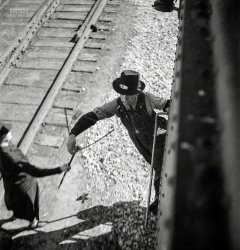 March 1943. "Dalies, New Mexico. Conductor C.W. Tevis picking up a message from a woman operator on the Atchison, Topeka &amp; Santa Fe between Belen and Gallup." Photo by Jack Delano for the Office of War Information. View full size.
Clarence W TevisFound in the 1940 census with wife Ferol? in Gallup New Mexico. Both born circa 1891. Listed as Railroad Conductor. Died 4 June 1971 in San Diego CA. RIP 
PanacheExtraordinary photo, certainly shoots way up immediately in my Delano favorites.  Here's a man, not at all young, with strong and hardened hands, performing a physical task nimbly and efficiently -- while smoking! -- with a cigarette holder!!  I look forward to the comments on the details of this message device.  (The woman: a blurred statue beside the dynamic conductor, caught crisply at precisely the right moment.)
Re: Cigarette Holder.In those days, a lot of men rolled their own smokes as it was far cheaper than buying premade. Usually, this led to loose tobacco getting into your mouth, on your face or clothes (been there, done that). As a result, many chose to use a cigarette holder to crimp the smoke and keep the tobacco where it belonged. It wasn't just the fashion statement as in the case of FDR!
The message device is called a Train Order Hoop even though it is Y-shaped.  The name comes from the shape of an earlier device that was used for the same purpose, to deliver messages to non-stop trains as they passed a station.
The paper containing the message was tied to a loop of string that in turn was held by the 'hoop'.  The man on the train would stick his arm through the loop and snag the string with the attached message.
This was a improvement over the older system where the entire hoop was snagged.  After the message was removed the hoop was thrown from the train for the person on the ground to retrieve, sometimes quite a distance down the track.
(It wasn't a great feat to get a crisp picture of the conductor, he was traveling at the same speed as the camera.) 
The practice continued This practice continued on Class ones until the advent of cab signals. Here is a Conrail train picking up orders at a temporary block station in November, 1978
On message hoops..There were variations - we at the CPR used a steamed wood hoop design, made in Angus shops. These worked well, unless you were the station junior clerk who had to gather them up from down the line after they were dropped by the train crew..in the pic, notice the flimsy dates from the Multimark era (the Multimark was in use from 1968 until 1987 or so) 
A. V. O.The "flimsies," so called because of the lightweight paper used, contained dispatching orders for the train. For example, that they should proceed to siding xxxx, clear the main line, and wait until train number YY passed before proceeding. This was part of an elaborate system of decentralized traffic control, documented in a book called "Rights of Trains," revised by Peter Josserand, head dispatcher of the Western Pacific railroad and a friend of my father. Flimsies and other forms used by the WP typically carried the letters "A. V. O." at the top, which stood for "Avoid Verbal Orders." Misunderstandings could be fatal.
Why &quot;Flimsies&quot;Some of the other commenters have mentioned that the old-time train orders were nicknamed "flimsies" because they were on lightweight paper.
The thin paper allows light to come THROUGH the paper. This allows the order to be read by the light of a dim kerosene lantern or even the light of an open steam locomotive firebox door.
This practice of using translucent paper continued far into the diesel locomotive era. Bright interior lighting is not wanted in any locomotive cab - it cuts the crew's night vision.
TimingDoes anyone know how fast this train would have been moving?  I know nothing of railroads, but quite a bit about photography, and I'll say that even with great skill, the perfect timing of this exposure involves at least a little bit of luck for the photographer.  And the faster the train was moving, the greater the luck/skill ratio required.  Until a definitive answer arrives, I'll give an educated guess based on the relatively limited motion artifact that the train was not going very fast at all.
More about &quot;Flimsies&quot;In addition to what SouthBendModel34 said, the paper used was thin to make it easier for the agent or operator to write multiple copies "in Manifold".
Double sided carbon paper was used and placed behind the first page of a manifold, and behind each of the other odd numbered pages.  If handwritten, a stylus was used as a writing instrument - not a pen or pencil.  If typewritten, typewriters without ribbon were used.  The first page and all subsequent odd pages had the message on the backside of the paper, and were read through the paper.  Even numbered pages had the message on the front.
As many as 10 pages could be prepared simultaneously, whereas if single sided carbon paper was used only half that number could be prepared at once.
Another feature of the "flimsies" paper is that is was fairly waterproof, and that messages from the carbon paper did not smear.
Here's a 60 year old example of such a flimsie as might be handed up to a crew as shown in the original photo.
(The Gallery, Jack Delano, Railroads)