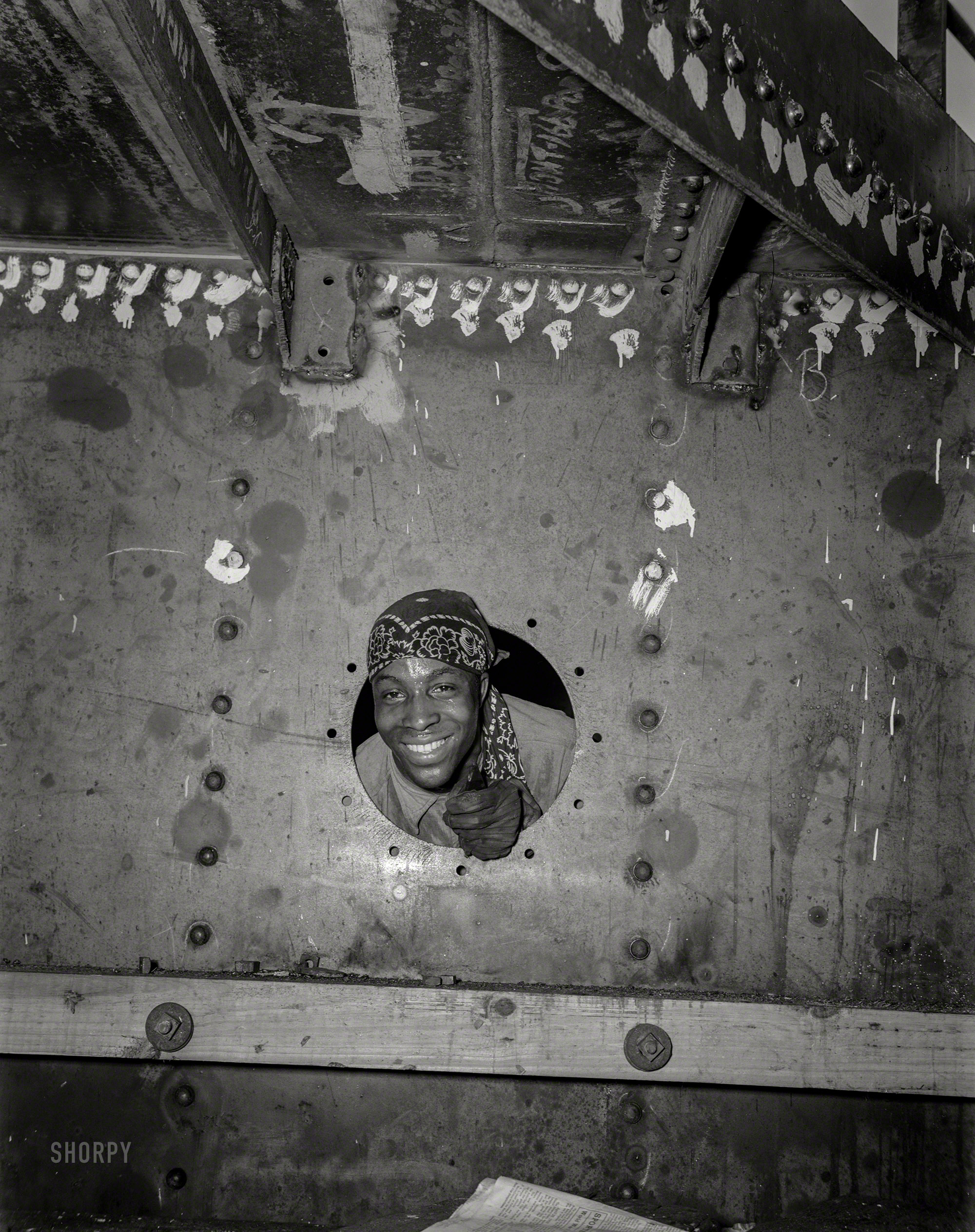 May 1943. "Baltimore, Maryland. Building the SS Frederick Douglass. More than 6,000 Negro shipyard workers are employed at the Bethlehem-Fairfield shipyards, where this Liberty ship is being rushed to completion. Douglass, the noted orator and abolitionist leader, worked as a ship caulker in the vicinity of this yard before he escaped from slavery. Smiling from porthole of the dockhouse is rivet heater Willie Smith." 4x5 inch nitrate negative by Roger Smith for the Office of War Information. View full size.