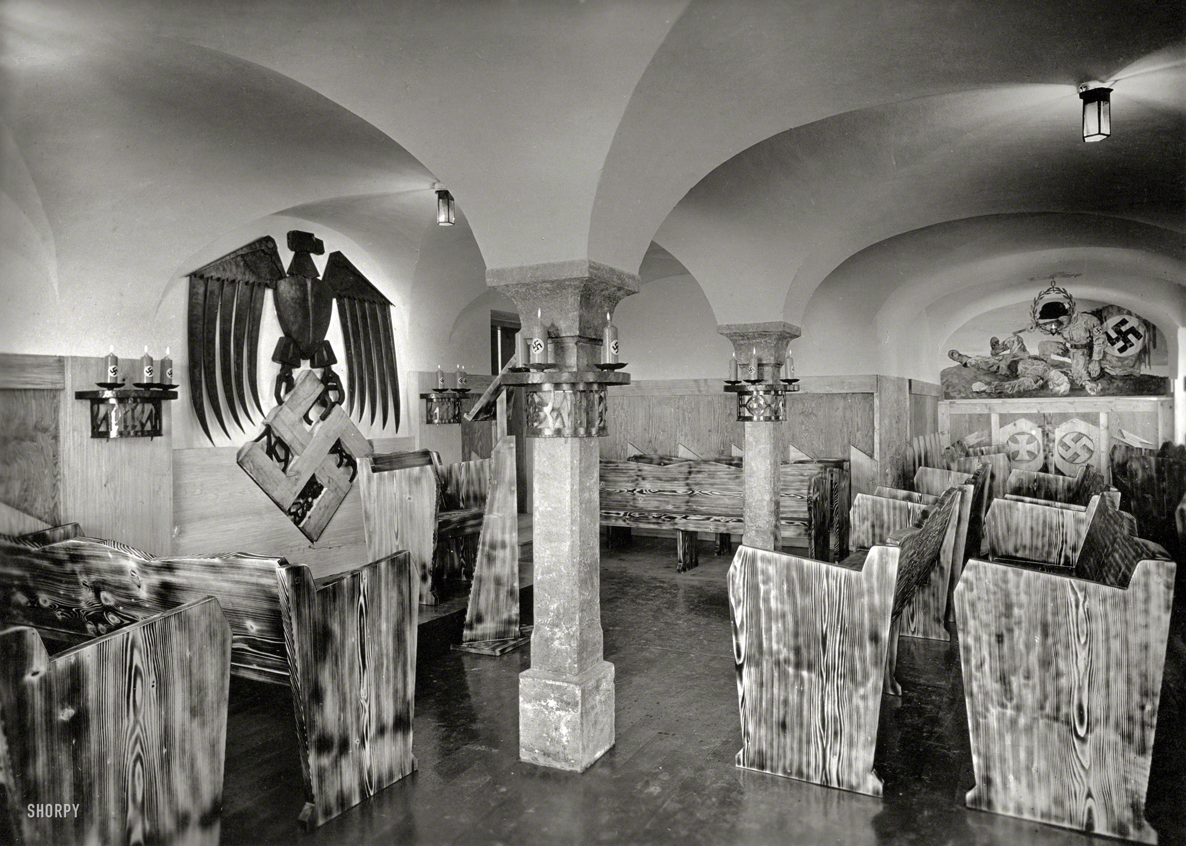 Germany circa 1938. "Meeting room of Nazi Party facility in Upper Bavaria." The swastika candles lend a rustic note. This looks like something out of a comic book or movie serial, but it was all too real. Photo-Pfaller print. View full size.

Additional details on this photo, one of seven pictures in a portfolio that's part of the Third Reich Collection at the Library of Congress:

One portfolio (seven photographic prints); 13 x 18 cm. Photographs on mounts 23 x 29 cm. No captions. Ink stamp on back of mounts: "Photo-Pfaller, Traunstein/Obb. Tel. 451." Confiscated by U.S. military intelligence authorities, 1945-1946. Transfer; 1947. Photographs show a chapel (?) in a Nazi party house in Bavaria. Includes wooden carved benches and podium; elaborate Nazi eagle and swastika symbol made of wood; mural of fallen soldier and SA companion; light fixture incorporating a helmet. Also includes exterior views of the house showing a mural with SA soldier with swastika flag and farmers with tools.