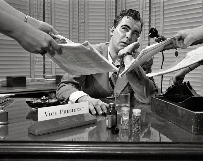 June 1952. Cornered in his corner office:  "Harried executive sitting at desk with pill bottles  and ashtray full of cigarette butts; arms are handing him papers and a telephone." Photo by Phillip Harrington for the Look magazine assignment "Your Next Promotion Can Kill You." View full size.
