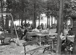 1925. "Glen Echo Park Co." Picnic tables at the Montgomery County, Maryland, amusement park. National Photo Company glass negative. View full size.
Picnic groveThis looks like the same area, near the bumper cars, where I waited for several hours around 1955 to meet Buffalo Bob from Howdy Doody.
After the long wait we were rewarded with Buffalo Joe or some such name as Bob had a more pressing commitment. 
AmenLet's eat!
Suitable attireI'm surprised to see so many men in suits. It doesn't look like a lot of people having fun. I wonder if was a company picnic. And of course, everyone at Glen Echo was white, and would be until 1960.
The Cloche Hatsitting on the picnic hamper in the immediate foreground would look perfect on the bobbed haired flapper standing left of the tree!
So GladThat Glen Echo still exists and that some of the rides have been restored so that everyone can enjoy them.  This was out in the 'country' when this picture was taken and a street car line carried you to the Park from Georgetown.  
American Beer GardenThe outdoor atmosphere, the trees and the picnic tables hark back to the German-American beer gardens that were once common.  However, based upon the somewhat sad to uninterested faces, like the man in left center, it's clear this is indeed during the less than Gemutlichkeit period of American Prohibition.
Post more!I have fond memories of Glen Echo from my childhood. I can still see the airplane ride at the entrance to the park. Please post more pictures!
And yes, it was all-white. Washington DC was, alas, much segregated until the mid-1960s. The Maryland suburbs weren't much better, and Virginia was simply shameful. I myself grew up listening to kids casually using the "n-" bomb. 
Still ThereThat area is still full of picnic tables. Although its no longer an amusement park, its still a great place to visit. The Bumper Car Pavilion is used for dancing (real dancing, like swing) in the spring and summer seasons and the glorious Spanish Ballroom from the 1930's (where my grandparents danced) holds dances all seasons. I've gone swing dancing there and it is magical.
(The Gallery, Natl Photo)