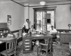 Washington, D.C., 1920. "Faulkner Inc. -- office interior." And a well-equipped office it is. National Photo Company Collection glass negative. View full size.
Summertime bluesAugust in D.C., no air conditioning, ties, and starched collars. My idea of the inner circle of hell.
At least they got to roll up their shirtsleeves.
It&#039;s For You Mr FaulknerPlease pick up the extension phone.
Casual Friday?Where are their suit coats?  Shocking.  And rolled up sleeves?  
Can't wait for other Shorpy comments to explain that device on the central desk.
Phone on pantograph!Mighty masculine place. Even the calendar shows a male model.
I have never seen a stick telephone on a pantograph extension arm before. Interesting! Now I'll probably see many more, so it must have just been invented.
Ergonomic It&#039;s NotI would hate to have to sit in one of those wooden chairs all day long. A partner desk with someone across from me is definitely not on my list. 
Curious ContraptionThe odd looking device on the desk is either a scale model of the 20th Century Limited or a Protectograph Check Writer.
Desk holes: early Schellenberg design?  Walter Schellenberg was a very senior SS officer doing international spy work for the Nazis. He was infamous for his "office fortress" desk, which had two automatic guns built into it that could be fired by the touch of a button
The extension arm forThe extension arm for desk-set telephones had been around for at least 20 years and they are quite often for sale on EBay. It was ubiquitous from 1905-1925 when the last incarnation was extension with a small square table for the newer Kellogg Grabaphone and Western Electric model 102 integrated handset and later telephones into the 1940's. Here is a short discussion: http://www.classicrotaryphones.com/forum/index.php?topic=5053.0
That Devil-May-Care VibeYou can't tell me these three jaspers don't get real crazy at the annual Christmas bash. And the gals over at Accounting? Don't even ask! 
Wall calendarCool calendar on the wall.  You have three consecutive months visible at all times, and you tear all three off on the first of the month.  Clever.
All that old fashioned office stuff, desks, stamps, heavy glass containers for whatever, they're all such neat artifacts.  Think how many millions of the grand old oak desks got tossed into the dump when the steel ones came along.
Balcony, or long drop?Judging from the building next door, this is at least on the second floor, and the if the window or french door opens onto a balcony or fire escape, I see no railing.
Better hold that Christmas party downstairs.
You got me...The boss guy looks like he took a bullet or something.  Anyone have a better idea?
Stick telephone on a pantograph extension You have seen one luojudson. It fugured in a Three Stooges short where the boys ended up dangling from one out of the window of an upper floor office.
Just the facts, ma&#039;amHoused in the Munsey Building at 1329 E Street, N.W., on publishers' row, Faulkner Inc. published The Faulkner Reference Library ("Standardized statistics of the United States"), and the Space Buyers Reference Library ("Pages of dependable facts condensed into periods").  The latter received at least one rave review, by 1920s standards.
16 AAAsDapper Dan at the desk has got the longest, narrowest and highest arched feet I've ever seen.  They must have been custom-made by elves.  The shoes, not the feet.
Who&#039;s the boss?The guys with all the authority were the ones who possessed the rubber stamps.  One used to know one had "arrived" when he was in control of the rubber stamps.
(The Gallery, D.C., Natl Photo, The Office)