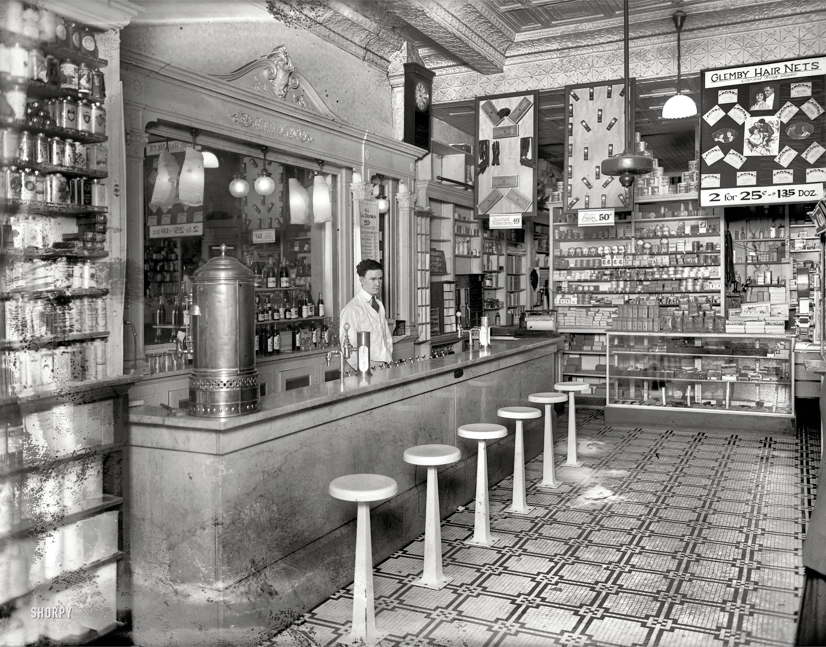 Washington, D.C., circa 1920. "People's Drug Store, 14th & U Streets, interior."

Is flyaway hair holding you back? Keeping you home those evenings your friends are out having a gay night on the town? Those loose strands may be a symptom of deeper ills -- malnutrition the consequence of egg-based-beverage deficiency, perhaps aggravated by tooth-brushing with an inferior dentifrice. Ladies, am I talking to you? Then come see this man. He is the gatekeeper to hair-nets, toothpaste, Egg Drinks -- and so much more. (Rubber gloves, for one thing, but that's another story.) The day you pick up a Glemby Hair Net -- or two, or hell, why not a dozen -- is the first day of the rest of your life! View full size.