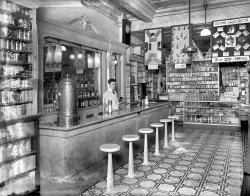 Washington, D.C., circa 1920. "People's Drug Store, 14th &amp; U Streets, interior."
Is flyaway hair holding you back? Keeping you home those evenings your friends are out having a gay night on the town? Those loose strands may be a symptom of deeper ills -- malnutrition the consequence of egg-based-beverage deficiency, perhaps aggravated by tooth-brushing with an inferior dentifrice. Ladies, am I talking to you? Then come see this man. He is the gatekeeper to hair-nets, toothpaste, Egg Drinks -- and so much more. (Rubber gloves, for one thing, but that's another story.) The day you pick up a Glemby Hair Net -- or two, or hell, why not a dozen -- is the first day of the rest of your life! View full size.
Hard Uncomfortable SeatsApparently counter space is at a premium and the stools here were installed to ensure a rapid turnover. Speaking of seats, if a physician ever approached me wearing one of those rubber gloves there would be a fight.
Oh boy!I'll have a Cherry Smash!
Molecular physicsI was a soda jerk one summer in the '50s. One day the boss handed me a large bag of sugar and pointed to a full bottle of water, and said "Mix it in." I complained that there wasn't enough room for all that sugar. He just smiled and said "Try it." A half-hour later the sugar was all gone, the water level hadn't risen perceptibly, and I had a jug of simple syrup.
The drug store was Easterling's in Jacksonville, in case there are any other natives out there.
Bob was confident, so very, very confident.Bob knew he looked darn swell, with that tire knotted just right by his mom, and the way his soda jerk jacket fit, and he was confident that, one day, when SHE came through the door, brunette tresses flying and probably a lavender silk scarf or it might be powder blue trailing behind, the scent of her alluring perfume trying to keep up, that she would see him there, and she would stop, suddenly. Yes, she would do that, because it was a very small store and if she did not, she would bump into the counter, maybe trip over one of those stools, and hurt her knee or even a foot. But he was ready for her. He could handle that, for he knew exactly which shelf held the mercurachrome.         
What is itThe big cylindrical gizmo with a spigot -- coffee machine?
Key PersonWho has the job of winding the clock?
Glemby Hair NetThe Lorraine Hair Net, manufactured by Glemby and made with real human hair, seems to bear a striking resemblance to a merkin!
Mr. Glemby: Rich Man, Poor ManAttached is an article from the January 22, 1932 edition of the NY Daily Star. Evidently there was good money to be made in hair nets.
It is somehow oddly ironic that Mr. Glemby's first name was "Harry"!
Coffee/tea urnHere's one that looks just like this one.
The jerkWhy is he doing nothing when there's all that sugar on the floor?
What&#039;s the deal.......with the covers (?) on some of the light fixtures ?
And wouldn't the fan work better if someone attached some blades to it?
Pre Cell PhoneIf I had a nickel, maybe I'd make a call on that phone in the back right corner.  As with so many of these photos, I'm crazy for the tin ceiling.
Barber shops and drug storesI remember that floor tile pattern in the 50's and 60's still in use even in public bathrooms 
Those bottles behind the young man might be flavors for the ice cream sodas and sundaes. Does anyone know for sure?
Soda Jerks&#039; ExposéLike Bull_Durham below, I was a soda jerk 50 years ago, too. I did my stent at a Jahn's Ice Cream Parlor in Coral Gables, Fl. There's a little secret amongst us jerks that I'll let you in on: there's NO egg in a "Egg Cream Soda". There isn't even any cream; it's just syrup shot with seltzer to make it foam up like meringue.
Sorry, not a lot a food value, but a real money maker for Mr. Jahn!    
Fascinating.Wonder how many of the medicines actually worked, and how many of those that worked can still be found on shelves or in the pharmacy today.
Only the finest ingredientsBehind our intrepid counterman are bottles of Clicquot Club, a popular brand of late 19th- and early 20th-century soda. For background, see http://en.wikipedia.org/wiki/Clicquot_Club_Company
What is thatWhat is that gizmo on the right wall?  Our soda man has a problem I see may times in shorpy, how to make bushy curly hair look glued down 20's style.  Imagine all that hair oil on a hot day.  
Site of the Start of the 1968 DC riotsFor some reason the Washington D.C. 14th and U addy rang a bell in this old man's head and the ringing proved to be correct ...
I am trying to imagine the scene 48ish years later when this store was the starting point of the 1968 DC riots.
http://www.washingtonian.com/articles/people/people-were-out-of-control-...
IHAP
Speaking of hair netsHere I am in 1974 in the main plant of Hershey with hair-netted Shirley Temple Black who’s learning all about making chocolate from someone who didn’t know much more about it than she did. She had just been named U.S. ambassador to Ghana, a major cocoa bean source. I couldn’t tap dance, either.  For high fashion fans, I am sporting a lovely light blue, white-stitched, denim suit with tastefully flared trousers and a dark denim belt. Note how my gorgeous paisley shirt picks up its pattern cues from Mrs. Black’s frock. Or the other way around.
The hairnet? Employees and visitors both had to wear them.  This was after public tours were ended and only VIPs were allowed (the massive production area was not designed for large numbers of outsiders who affected the factory heat and humidity levels, and there might have been safety concerns).  Few people can say, as I can, that I saw the beautiful Dorothy Hamill wearing a Hershey’s hair net. Glemby or not, she still looked darn good.  
What that isIs the end of the cash register.  What we're seeing is the printing mechanism and the paper tape.
Egg CreamsAs a resident of Brooklyn and not of the Sixth Borough - South Florida, with all due respect to bryhams...while (s)he is correct about egg creams having neither egg nor cream, it does have milk. Without the milk, it's merely a chocolate soda.  And, for it to be a TRUE egg cream, it has to be made with U-Bet chocolate syrup.
Stool sampleSame stool bases as Boo Koo burgers down Texas way!
https://www.shorpy.com/node/12857
(The Gallery, D.C., Natl Photo, Stores & Markets)
