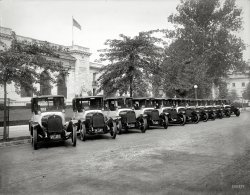 Washington, D.C., 1922. "Black &amp; White taxis at Pan American Union." National Photo Company Collection glass negative. View full size.
What make are the taxi cabs?Anybody have a guess?
Interstate CommerceWere those taxis allowed to drive customers to either Maryland or Virginia? I sort of remember seeing dual Md and DC plates on autos in photos of that era.
Built by the Yellow Cab Manufacturing Co.As founded by John Daniel Hertz. More on this company here...
Black &amp; White &amp; YellowThese cabs were made by the Yellow Cab Manufacturing Company of Chicago which was owned by John Hertz.  The logo on the radiator looks almost exactly like the pin shown below.  Another view of the cab is shown as well.
For Washington, D.C. to not require cabs to have true headlights in 1922, as compared to the cowl lights shown, seems strange.  Most major municipalities, and certainly almost every state, had laws mandating required motor vehicle equipment by this time.  Even worse, the cowl lights of Black &amp; White Cab were not clear glass.  Many also had the company name placed in them further reducing their ability to project light.  An example of this is also shown below.    
John Hertz is who Hertz Rental Car is named after, and the yellow Hertz logo harkens back to the days when he owned both Hertz and Yellow Cab.
The Pan American Union Building is on 17th Street NW between C Street NW and Constitution Avenue NW.  It took almost two years to complete and was dedicated on April 26, 1910.
Headlamps optionalNone of the cabs had expensive and unnecessary headlamps installed. After all, these are work vehicles. But the cowl lamps were electric units.
Disorderly spotlightThe guy on the left doesn't have his spotlight aimed correctly.  Darned useful things, have often wished my present car had one of those.
(The Gallery, Cars, Trucks, Buses, D.C., Natl Photo)
