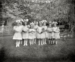 July 22, 1913. Washington, D.C. "Dorothy's party." At center is Dorothy French, daughter of National Photo Co. proprietor Herbert French. View full size.
Left OutThe poor little girl on the other side of the fence looks so forlorn.
Did the invitation specify a dress code?What a sweet group.  I bet the solemn faces disappeared when the cake and ice cream arrived!
BackgroundThere's a neighbor maid's daughter on the other side of the fence.
Not serious enoughYou - You there in the back!
Wipe that "smile" off your face!
Flappers!The fashion for big, floppy hair bows for little girls in the 'teens lead to the name those girls would go by as they became young women in the 'twenties - flappers. 
Sock CorsagesThe little girl with the colorfully dressed doll (the birthday girl, probably) has rosettes at the top front of her socks. I don't think I've ever seen anything like that. VERY cool!
High button shoesMy Mom grew up in Brooklyn.  She used to tell me she hated the high button shoes they had to wear.  Some of these girls are wearing Mary Janes, which look like shoes worn today.  None of these girls look too happy but it's a great photo.
Little girl lostI would like the think that they would have preferred to forget the photo so they could play with the little girl on the other side of fence. 
(The Gallery, D.C., Kids, Natl Photo)