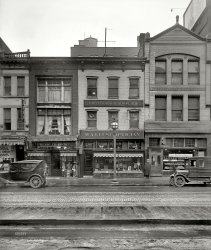 Washington, D.C., circa 1926. "M.A. Leese Optician, 9th Street store." Other businesses represented in this mold-spotted street view include a pool hall, Venice Italian American Restaurant, Pressler Bros. haberdashery and Barker "Original System" Bakery. National Photo Company glass negative. View full size.
Beating the dead hatDave said, "If it was the same guy, we'd be able to see through both of his images and not just one. The door behind Mr. Left would register at least as strongly (or faintly) as the body of Mr. Right."
The transparency of the man would be proportional to how long he was in each position. He was standing at the left for a longer time than he was standing on the right. That Mr. Left was in motion I don't think is in doubt; if you look at the close-ups below, you can clearly see the sidewalk through his legs and feet. His hat also has a clearly visible trail as he walks to the right. I think you can also see the line of the building through his right arm.
[By the same reasoning, the apparentness of the building is proportional to how long it's in each position. Thinking solely in terms of how transparent the man is is misleading; the important thing is the relative prominence of each element in each man-building pair. If it's the same guy, the building behind the man on the left would be at least as prominent as the man in front of the building on the right -- if he was standing to the right for 5 seconds, this means the wall behind the man on the left has been exposed to the camera for 5 seconds, plus the time it took him to get from A to B. However this is clearly not the case -- although it's obvious that Mr. Left has moved, the building behind him has been exposed to the camera for just a fraction of the time that Mr. Right has been standing in front of the window. To summarize: The relative prominence of the fainter element of each of the two man-building pairs should be the same if it's the same person. On the right there is very strong evidence of a man in front of the building; on the left, however, there's hardly any trace of the building behind the man. - Dave]
A radio pioneerM.A. Leese established one of Washington's early radio stations, WMAL, whose name incorporated his initials. Today at 630 on the dial, it is the home of right-wing talkers.
[Note the antenna on the roof. - Dave]
Very long exposureThe exposure on this one was so long that the man in the picture had time to turn back around, walk 3 or 4 paces, and then stop to look at the bakery window.
[Those are two people; different hats.]
Hmm. I'm willing to be convinced, but the two men have the same height, the same coat length, the same hands in pockets, the same ruffled sleeves, and the same shades of gray. I see a scarf around Man #1 and a dark spot on the back of the neck of Man #2. The hats are each light colored with a dark stripe. The shoes and pant legs are obscured in Man #2, but is still consistent.
The only inconsistent thing I can see about the hats is that the brim of Man #1 seems upturned and the brim on man #2 seems perhaps too wide and flat. But it could just be the angle and blur of the photo.
His Hat Brimmeth OverIt appears to be raining. The mystery man may have exited the building with hat brim turned up; "click" - first exposure; seen the rain; flattened his brim for maximum rain protection before stepping off; put his hands back in pocket; stepped off; "click" - exposure two. All the while quite unaware of his future stardom on Shorpy, and all the controversy he would cause!
Which Should I BringThat "POOL" sign over the soon to die jewelry store makes me wonder whether I need my pool cue or my swim trunks.
AccessHow did one reach the Italian restaurant, through the haberdasher's store or through the pool hall. 
[Take a close look at the passageway next to the jewelry store.]
But isn't that the 'next' building over?  They must be connected above the first floor.  Thanks
Re: Checking to see if the coast is clear@Quatermass -- You do see a smear... note on the right side of his hat, you see the motion blur, which is stronger while he's turning and starting (less motion) and fades out as he accelerates into his walk.
I see Dave is sticking to his story of the different hat, despite my carefully crafted evidence of everything else being the same (including 90% of the hat).
Kidding aside, I think the coat sleeves are the killer evidence. Besides the identical wrinkly nature, if you look at the right arm, there is an identical "puff" at the elbow.
Thinking about the hat one more time, maybe what I see that Dave doesn't see (or doesn't agree with) is that the top round part of the hat is over the ship on that white sign, obscuring it, making it look like a different hat. If you look carefully, you can see the ghost of the top of his hat where the color is slightly darkened over the sign. I see the same round top with dark band. See pic below, that I created from the original LOC image at max resolution.
[That's not Dave who was taking issue with the comment below. That was our new Comments Czar. As for whether it's one or two people, the hat is beside the point. If it was the same guy, we'd be able to see through both of his images and not just one. The door behind Mr. Left would register at least as strongly (or faintly) as the body of Mr. Right. - Dave]
Antenna sizeMaybe with just 15 watts 15 watts it took that much of an antenna to get them even at a short range (not certain how far apart this store and the transmitter site were then).  The article notes the numerous changes in frequency that many stations went thru in the 1920s as broadcasting developed.
Checking to see if the coast is clear@ TimB: I'm pretty sure that's the same guy.  It would have to be a double exposure though, or else we'd probably see a smear of him walking over there.
My idea was that he had to look around to make sure the wife wasn't watching, then quickly step over to the bakery to purchase a clandestine eclair.
[But first stepped down the street to buy a new hat.]
Okay, so maybe one of them bought the eclairs and the other is the lookout.   
Come to think of it, that might be Herman and Clarence Woolard, of the famed Jelly Roll Gang, who terrorized bakeries up and down the Eastern Seaboard throughout the 1920s.  Though history has tended to recall the fanciful exploits of gangsters like Dillinger and Bonnie and Clyde, it's said the Woolard's made off with a lot more dough.
Vision DeteriorationThe mold makes the three windows of Leese's building look like stages of some horrible degenerative eye disease.
The eyes have itFor those not sure what an optician does just take a  look in the windows.
Enough already!I'll call Old Man Barker and ask him who was hanging out in front of his shop that day. Sheesh you guys!
Old Man BarkerProbably wasn't at this bakery.  The Barker Original System of Bakeries was a sort of franchise, where the manufacturer of the baking equipment would set up a new baker for an investment which naturally included buying their equipment.
Advertisement in the Pittsburgh Gazette Times, June 22, 1919:
(The Gallery, D.C., Natl Photo, Stores & Markets)