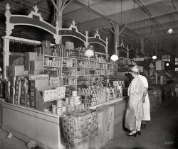Washington, D.C., circa 1926. "Thomas R. Shipp Co. -- S.A. Gatti stand, Center Market." Offering malt extract, wine jelly and hops, three items for your Prohibition-era shopping list. National Photo glass negative. View full size.
LoopholeI believe that during Prohibition it remained legal to make beer or wine at home for family consumption (though not for sale or other distribution).  Distilled spirits remained outside that exemption, however.
Still Going Strong!Among the hops and malt are other product names that have survived and are still recognizable brands: SOS - scouring pads, Libby's - vegetables, Angostura - bitters, Crisco - shortening. Household staples still!
And the Winner of the Blue Ribbon isWhen I was still in highschool I had a job at a local IGA grocery store. They sold Hop Flavored Blue Ribbon Malt Extract. I wondered what good anything could be that was flavored by hops. Then I learned how to make home brew. My question was answered.
MaltWhen I moved to Georgia in the 60's it was exceedingly hard to find any alcoholic beverages, much less pizza. Necessity being the mother of somebody I was able to obtain an old 5 gallon glass water jug into which I placed a 3 lb can of Blue Ribbon Malt and 5 lbs of sugar, topping up same with water - which in a week or two made an almost serviceable beer, although on occasion the stuff would would overflow the jug making a bit of a mess.
Strange posterWhat is that a poster for just to the left of the womans hat brim?
I can't zoom in on it enough to see.
[Bertozzi Parmigiano Reggiano cheese. - tterrace]
Hot tin roof (but no cat in sight)I have noticed that in many of the pictures of stores on Shorpy, they appear to have some type of corrugated metal ceiling.  I would have to hazard a guess this must be a lower level from a multi-story building, as it doesn't look sturdy enough to support the occasional heavy winter snow that might come to the DC area; and there also doesn't appear to be any insulation.
[Photo of the exterior here, along with a link in the comments to more photos inside. -tterrace]
Re: Strange PosterThere's another Bertozzi poster to the left of the "cheesy" one:
Gatti Bros.


Washington Post, April 30, 1961.

Seraphin A. Gatti Dies; D.C. Wholesale Grocer.


Seraphin A. Gatti, 80, a wholesale grocer in the District many years, died of heart ailment Friday in Arlington Hospital.

He was one of the first institutional wholesale grocers in Washington, serving restaurants, hospitals, schools, clubs and government establishments.

Mr. Gatti started in the retail business shortly after arriving here in 1886 from his native Italy. He ran a grocery with his brother on the site of the Archives Building. Later he went into wholesale work with his son, and retired in 1952. &hellip;




Washington Post, December 20, 1969.

Louis B. Gatti Dies, Produce Merchant.


Louis B. Gatti, a retired produce merchant whose fruit and vegetable stand won the culinary cachet of early 20th century Washington, died Thursday in Georgetown University Hospital after a long illness, He was 96.

His stand was one of many in the old Center Market, a two-block wholesale and retail produce emporium located on the present site of the National Archives in downtown Washington.

But the fine quality of his vegetables and fruit&mdash;such as strawberries from Plant City, Fla., and typhoid-free watercress grown in pure mineral springs&mdash;won an elite clientele of the rich and powerful in a day when even the most haughty of the city's housewives did their own daily grocery shopping.

Perhaps his most discriminating customer was Mrs. William Howard Taft, wife of a gourmand, the 27th President of the United States. The day after her husband's election in 1908, she appeared at Gatti's stand to let him know that she would soon be taking delivery at the White House.

President Taft lost the next election but apparently Woodrow Wilson found Gatti's produce just as tasty. He continued to supply the White House, his wife Mary Angela, doing the selling, he the buying. &hellip;

(The Gallery, D.C., Natl Photo, Stores & Markets)