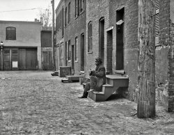 Update: This is the west side of Blagden Alley. Details in the comments here. "City rowhouses, 1923." Another view of back-alley Washington, D.C., and its long-forgotten habitues. Harris &amp; Ewing glass negative. View full size.
Can&#039;t make it outI wish I could make out what he is doing or repairing. It looks as if he might have a crochet hook in one hand.
But Clean!These DC alley shots are intended to convey poverty and perhaps despair, yet one is struck by how tidy these precincts are -- no piles of detritus, broken windows, or abandoned cars.
(The Gallery, D.C., Harris + Ewing)