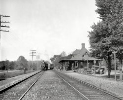 Short Hills, New Jersey, circa 1901. "Short Hills Station -- Delaware, Lackawanna &amp; Western R.R." 8x10 glass negative, Detroit Publishing Co. View full size.
Short HillsShort Hills was developed in 1879 by Stewart Hartshorne, who made a fortune on window shade rollers. At first, the residents used the nearby Millburn depot, but after the DL&amp;W promised to stop at least two trains here daily, Mr. Hartshorne donated the land and spent $2500 to build this station. He paid the agent's salary here and maintained the building at his own expense for a number of years.
Eventually, this depot was deeded to the Lackawanna, and served the town until 1907, when it was demolished to make room for its replacement. [This information was lifted from "Delaware, Lackawanna and Western Railroad in the Nineteenth Century" by Thomas Townsend Taber]
There is a fence nowbetween the tracks. I'm pretty sure that this view is the same direction from about where the conductor is standing on the left.
Pole lightsI have never seen the way the light shades on the poles are placed. It appears that they are upside down but I'm sure they are not. Beside that it looks like it is going to be a nice day as the men hang out watching the woman folk head out on the rear of the train, perhaps a shopping spree in NYC?
Flat LandscapeLooks like they're about one hill short.
Short Hills in 2007View from roughly the same angle, 106 years later:
Looking west (young man) not eastJudging from the sun angle and the orientation of the tracks (basically NW-W to SE-E), I think the picture is looking west towards Summit, NJ, the next stop on the line and the highest point on the Morris and Essex Railroad (builder of this line and purchased by the DL&amp;W prior to this photo).  The excellent photo posted by swaool is looking eastward towards Millburn.  If it is indeed looking west, it would make the station building on the westbound side of the tracks, which is a bit unusual since most passenger traffic waiting for a train was and is eastbound towards Hoboken/NYC.
I lived in Short Hills for thirteen years, and took the train to points both east and west many, many times from this spot.  111 years later Short Hills is a bit more built up than seen here, but is still a very pleasant community to live, and yes, it does contain many "short" hills.
(The Gallery, DPC, Railroads)