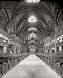 Circa 1900. "Main altar, Church of Notre Dame, Montreal, Quebec." 8x10 inch dry plate glass negative, Detroit Publishing Company. View full size.
Masterpiece!As genuinely glorious as Montreal's Notre Dame is, in this case I'm referring to the Detroit Publishing Company's wonderfully detailed photo of the basilica's interior. I thought it would be pretty easy to find a comparable modern color photo, and was surprised to find that, of the dozens of photos available in Google Images, many of them taken by professional architectural photographers, almost none of the images comprised as complete and undistorted a record of the Basilica's interior features as Detroit's carefully adjusted view camera version. It is of course exciting  but not surprising to learn that the primary color scheme is blue and gold. The Basilica's own website includes many fine detail images of the building's artworks and decorations, available at www.basiliquenotredame.ca/en/basilica/pictures.aspx, but no single interior view as technically accomplished as Detroit's. I did find one view online, despite its monitor-friendly horizontal format, that seems overall to come closest to the qualities of the Detroit image, taken in 2011 with a Nikon D90 by professional photographer Ash Henderson.
The OrganThe Organ can be heard and the present day interior viewed by clicking here.
A challenge to the coloristThis is one time when black and white really fails to do justice. Except for the pews and the central sculptures on the reredos and on the pulpit, virtually everything in this picture is either gilded or polychromed, including the ceiling. The pillars are polychromed and gilt in patterns. And they weren't finished, even then: the stained glass didn't arrive until the 1920s, and at some point someone apparently felt that the one blank area on the back wall of the apse above the reredos needed some decoration too, so they put a sort of celestial background on it-- gilded, of course. The replaced the altar with a different one which incorporates three reliquaries; need I say, gilt over every square inch of their Gothic revival surfaces?
I have to say, though, that Gothic revival skylights are something only the Victorians could have though of. And alas, the light fixtures among the pews are no longer with us.
Notre Dame Basilica, MontrealGreat interior view -- would love to see a shot from the opposite direction of the Casavant organ (1891) in the rear gallery.  (I'm an organist.)
Pipe questionThose sure do look like pipes along both sides of the aisle. Could they be for heat? Hot water pumped from a boiler? 
Similar PhotosThis photo is reminiscent of https://www.shorpy.com/node/6456
PipeCan that be a pipe running along the floor next to the pews? So out of place if it is.
Well, to me it looks likethe fanciest zeppelin hangar in the world.
Be Careful Be careful when you compare this photo with the modern views. The church was heavily damaged by fire in 1978, and most of the present interior dates from after the fire.
Heat requiredAs a Montreal resident I can certainly attest to the need for heat, it was 10 degrees F this morning. Pipes of this type are very much associated with hot water heating and the proximity to the rows of pews, along with the unusual detail of doors to the pews which clear the pipes suggests that these pipes may have fed arrays of pipes that passed under the foot space in the seating areas which, with the little doors closed to block drafts, may have provided sufficiant heat to enable parishioners on a cold winters day (-10F is not unusual) to sit through the two hour long latin masses that were typical of the day. Just guessing though.
PipesA quick check with an older family member who used to frequent that cathedral a long time ago confirms that the pipes were for hot water heating, with radiators below the "prime" seats.
You'll notice that most of the pews in the central rows have a door and a little plaque.  That was because they were private seats for wealthy Montreal families.  A "generous" contribution to the parish got your family a private pew in a prime well heated location and a plaque with your family name for all to see.
Be careful of being carefulThe smaller Chapel of Notre-Dame du Sacré-Cœur was damaged by fire in 1978 and was rebuilt afterward, but the main basilica interior is still largely original. Or so I believe.
(The Gallery, DPC)