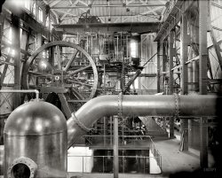 Copper production circa 1906. "12,000 horsepower compound pump, Calumet and Hecla stamp mill, Lake Linden, Michigan." Note the faint double exposure in this 8x10 inch glass plate. Detroit Publishing Company. View full size.
Enter Charlie ChaplinLooks like the movie Modern Times could have been shot there.
The Main Bearingmust have required constant attention as evidenced by the captain's chair on the platform. Employees of the era were rarely offered the opportunity to sit while on the job.
It brings to mind a John Lennon lyric from a 1980 song "I'm just sittin' here watching the wheels go round and round, I really love to watch them roll."
The steam engine is a work of art but so, too, is the building with its myriad girder and truss work. Note the sloping outer walls fit to the angle bracing and that huge overhead hoist! Another fascinating Shorpy view!
Sitting roomI found two other chairs, just to the right of the big wheel, one on the top platform and one on the bottom.
Domestic plumbingApparently they are pumping houses through that big pipe.
[The houses are an exact match for another Shorpy image -- who'll be the first to find it? - Dave]
The First Exposure or the Second?I propose The Heart of Copper Country: 1905 as the second exposure on the above photo.
[We have a winner. -tterrace]
Way ta go!Congrats to Orange56!  Good eye!
CraneCan anyone make out the manufactures sign on the Overhead crane?  Thanks in advance
(Technology, The Gallery, DPC, Mining)