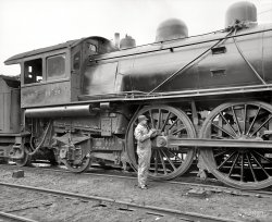 Circa 1904. "Michigan Central Railroad. Oiling up before the start." 8x10 inch dry plate glass negative, Detroit Publishing Company. View full size.
You railroad guysI love you railroad guys!  My dad was a machinist in the Northern Pacific shops in Livingston, Montana from the 1930s through 1950s.  I barely remember the steam locomotives, but I do remember when the diesel locomotives replaced them.  And I remember how my dad talked about the steam engines and their various parts and features as though they were his children -- just like you all do.  Thanks so much for making my Shorpy experience even better!
MissingAxle-box cover seems to be absent on tender leading wheelset!
Lube problemsNot sure what the fellow is oiling--grease cups for the main driving rod and driving pin are both sealed. Behind the fellow one sees the forward tender truck journal cover is missing entirely (not just pushed up in the open position) and the lagging is falling out. Much more squeaking to come. Some smoke too. Nice shot though of state-of-the-art passenger steam power of the time.   
I love trainsFantastic photo. 100+ years later that train is a marvel.
PerhapsThe photographer told this chap to "stand there and look busy while I take this picture so that, in 107 years, people will try and figure out what you're doing." And it worked.
Familiar FaceWe've seen this guy on Shorpy at least twice before, here and here. He has a very distinctive nose.
Repack me, please.For all the big-time railroading entailed in this passenger engine and her hoghead, the condition of the tender truck is out of character.
That is a Fox truck. The heavy assembled sideframes are typical, and were thought to be more robust than the common, and unreliable archbar trucks then in use since before the War Between the States. A lot of railroads used Fox patent trucks on tenders, cabooses and freight cars.
A period of use found the Fox trucks did not hold up well under heavy use, and were quickly removed from the railroad scene.
What suprises me is the journal box seen on that truck. There is no lid, critical to keep dust, sand (from the engine) and other debris out of the journal. The packing is hanging out of the box, indicating it will be serviced before we depart.
In a journal box, the axle, fitted with a large brass journal, rotates in the bearings while being bathed in lubricating oil. The packing, originally cotton waste, lays in the bottom of the box filled with oil. If the oil runs dry, the journal gets hot and starts burning. When it stops burning, watch out! Next thing is for the axle to separate from the truck, causing a ferocious wreck. A missing box lid would almost insure problems if any distance is involved.
These friction bearings date from the earliest days of railroading and were not banned from interchange service in the US until the 1970's, being replaced with roller bearings. (Steam passenger locomotives, the Michigan Central Railroad and the oiling hoghead above were already long gone...) 
Domes againThis is the third look at this well-shod guy oiling the engine. And yes, those bulges on top of the engine are (L-R) the auxiliary steam dome, the steam dome, and the sand dome. 
The rail lineThe condition of the fishplate on the railway line, with a missing bolt, the other bolts loose; and belled edges on the plate and rail, gives one great concern as to the generally poor maintenance level on the lines.
Oiling around. . . .He appears to be oiling the driving boxes for the rear diver axle. The driving boxes house the journal bearings and lubrication cellars. The function of the driving box is to allow the whole assembly to move vertically up and down in the frame as the suspension travels. Failure to lubricate these can result in excessive wear and even jamming of the driving box, rendering the suspension inoperable in that position. When I oil round the running fear on a steam locomotive, I always remember "if it moves, it needs lubrication somewhere." 
(The Gallery, DPC, Railroads)