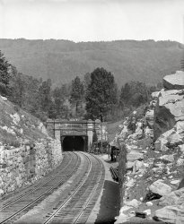 Circa 1907. "West portal, Hoosac Tunnel, North Adams, Massachusetts." 8x10 inch dry plate glass negative, Detroit Publishing Company. View full size.
Where&#039;s the garage door opener?Today the tunnel's west portal looks very different.  What's essentially a very large garage door was added around 60 years ago, though I'm not sure why.  The door's housing hides the "Hoosac" inscription seen in this picture.  The tunnel also has had a single track for many years, not the double track as shown here.
One remaining drawback is that the tunnel doesn't have sufficient clearance to handle double-stack containers.  There have been various engineering studies to see how that could be remedied, but the cost would be extremely high, and given that there's a more southerly route into Massachusetts with double-stack clearance it's doubtful anything will be done. 

Open and ShutWhy did they have doors on the entrance?  That seems unusual for a railroad tunnel.
That DoorI think that the Garage Door Opener (&amp; Closer) is partially sheltered in the nearest structure on the right.  It looks like there might be a pair of bi-fold doors and a wood track for them overhead.  The doors provide some protection from a "wind tunnel" effect that would allow cold winter air to defeat comparative warmth of being far underground.  Nowadays, in conjunction with intake and exhaust fans above the Central Air Shaft, they can regulate which end of the tunnel gets ventilated and try to keep the smoke away from the engineers' eyes.  Shortly after the Shorpy photo was taken, a short system of electric locomotives and wires was installed, to pull the steam trains through the mountain.  The coal smoke had gotten so bad that someone passed a red Stop Signal without seeing it!  Today, there are so few tracks and trains in the tunnel that it is hoped that the doors and fans can deal with the Diesel engine exhaust, and the electrics have been gone for 60 years or so.
Tunnel Doors"In 1954 a steel storm door was installed on the West Portal replacing the wooden doors. These doors helped keep strange weather from entering the tunnel, particularly in the winter. In 1957 The tunnel was reduced to a single track 3 feet north of the center for clearance purposes. On November 28th 1958 passenger service stopped. In 1973 the track was centered and replaced by continuous welded rail. Finally in 1997 a 10 foot wide strip of stone was removed from The Tunnel’s ceiling and the track was lowered to allow for even taller railcars. The rail at the East Portal was sunk below ground level."
from A Pinprick Of Light by Carl R Byron and Hoosac Tunnel History

The west portal circa 1874
Tunnel doorsEBT added doors to their tunnels after a locomotive derailed due to ice on rails in winter from ground water draining through tunnel. Doors were opened and closed by watchmen and later by engine crew.
Switch on; then offOverhead catenary wires for electric locomotives were added a few years after this photo.  The electric motors would couple onto steam powered trains and pull them through the tunnel so the steamers wouldn't have to work, thus creating a lot of smoke to asphyxiate the engine crews.  Even with ventilation shafts, tunnels were hellish in the steam age, and the Hoosac, almost five miles long, was one of the worst.  Diesel power eliminated the need for electrics, and the wires were then removed. 
Hoosac Tunnel: 1907About 16 years ago, I walked about 300 yards into the west portal. A fairly busy road passes only about a quarter mile from the entrance. Three or four times, I dashed out of the tunnel because I thought I heard a train coming, only to realize each time that I was hearing a truck go by. After about 15 minutes, I left, because not only was I pushing my luck regarding a train coming, but also because I was afraid I might startle an animal, such as a raccoon or a bear. I had my camera with me, so I decided to stand by the entrance, hoping that I might catch a shot of a train. I didn't have to wait more than three minutes. A train coming from the west (downtown North Adams) rumbled around the bend slowly and entered the tunnel, as I followed close behind, took a few pictures, and listened to the mysterious sound of the train gradually fading. Five miles later, it would see daylight again.
ElectrificationHoosac was electrified in 1910, and while Lost World is correct, Diesels did help, it did not totally do away with the problems crews faced.  Wasn't until the late fifties that things were markedly improved.
The tunnel was electrified after a number of crews suffocated in the tunnel itself.  It wasn't uncommon for crews to exit the tunnel lying on their stomachs in the cabs with cloths over their mouths and noses trying to get fresh air.  Even in the early days of diesel locomotives, there was still an ever present danger of this.  The Catenary didn't come down right away though, as the electrics were kept as backup until 1958 when the last one was finally scrapped.  Even so, much of the catenary was still in place (just disued) until 1973 when the tunnel was made single track.
West Portal DoorThis is the West Portal of one of my very favorite places, Hoosac Tunnel.  East Portal is way prettier.  The doors have something to do with keeping either water or snow out of the tunnel.  I know the exact explanation is in the book "Pinprick of Light".   The book also mentions failure of the brick lining on the west end due to (I believe) wetter conditions and far weaker rock (one reason why this end actually has a brick lining).  
Being a responsible railfan, I must add that this is NOT an abandoned tunnel, and while current traffic tends to be later in the day/night, a train can come by at any time.
 
(The Gallery, DPC, Railroads)