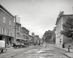 Circa 1908. "Main Street -- Littleton, New Hampshire." Yes, they have bananas, and Moxie, too. 8x10 glass negative, Detroit Publishing Co. View full size.
RE: MoxieFor anyone who is curious, Moxie tastes something like root beer with cherry cough syrup.
How did they do that?In 1908, how would you get bananas from Central America to inland New England before they spoiled? This little shop seems to have an ample supply.
I imagine that bananas were still a bit of a delicacy in 1908.
[USA annual per capita banana consumption in 1909: 19.3 pounds, according to the USDA. -tterrace]
Wires were different back thenIt's so strange to see the telephone wires above the power wires. There's a strict hierarchy these days of putting higher voltage wires above lower voltage wires. It's also strange to not see any high voltage lines or transformers on the poles. 
At least two survivorsThe building on the left with the columns and the sharply peaked roof is the well-known Thayers Inn.  It was almost 60 years old at the time of this picture and is still going strong today.  Also still extant is the Congregational Church whose steeple is visible in the distance on the left side of the street.
The far church is still thereAs are some left hand buildings in the background, starting with the flat face storefront with the four windows, and the inn behind it, with the tower, and four columns facing the street.
View Larger Map
Use and CareThe lady crossing the street wearing the long white dress obviously spotted a clear path to the other side. Rules of the day insisted on long skirts even though traversing a roadway could be hazardous to the well-being of the garment.
Thayers InnHere's a photo of the historic Thayers Inn I took while on vacation there in 2006. Established 1843, and glad to say it's still a very hopping hotel.
Bananas travel cool.The bananas probably sailed in United Fruit Co. steamships painted white to reflect the sun, and finally in railroad refrigerator cars from the port to the train station shown in earlier views of Littleton.
High voltageThe high voltage wires are there. The bottom crossarm carries the 110/220 household circuits, and three of the four larger insulators on the second crossarm carry the "high" voltage. It could be 1,100 volts, obsolete as soon as it was introduced in the 1890s, or it could be 2,200 volts, a system which was overwhelmingly common until the postwar era. The transformer is the horizontal black oval on the backside of the second pole. (If any transformers of this type are still in existence, there are collectors who will pay ridiculous amounts of money for one.)
The fourth wire on the second crossarm is for street lighting. Only one wire was needed, because it ran in a loop around the neighborhood, with the bulbs wired in series like Christmas lights. Nixiebunny is correct that running power below telephone would not meet current code. The reason it is like this, probably, is that the telephone wires were already in place when the power was installed.
MoxieMoxie, for anyone who has never tried it, is an experience.  Despite living in New England all my life I never acquired a taste for it.  I wonder if perhaps it tasted better in 1908 than it does today?
As for Thayer's Inn, it's a lovely old hotel. My husband and I stayed there around 2005.  We keep meaning to go back but haven't yet.  There are photos of Bette Davis all over the Inn because she lived in a nearby town in the 1930s (rumor has it she visited Thayer's quite often too) and one of her movie premieres took place at Thayer's.   
Moxie IIMoxie must have been good; Ted Williams endorsed it.
(The Gallery, DPC, Small Towns, Stores & Markets)