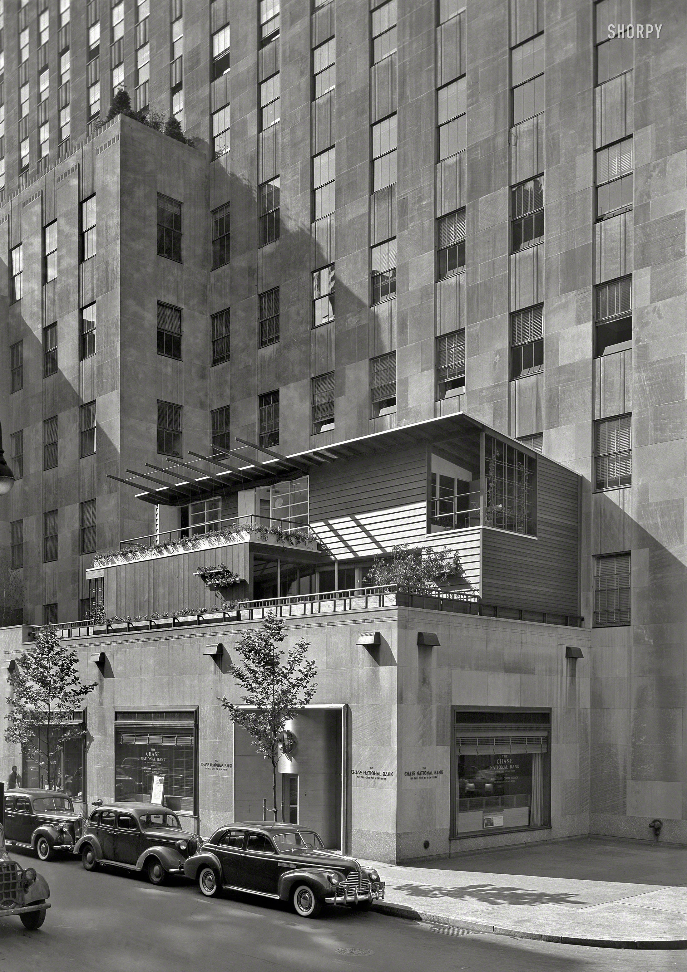 HOME EXHIBIT PLANNED
Dwelling Rises on Terrace in Rockefeller Center

&nbsp; &nbsp; &nbsp; &nbsp; NEW YORK -- On the second-floor terrace of the International Building at 51st Street near Fifth Avenue, a seven-room dwelling is under construction as the focal point in what is designed to be a home-building center and exhibit. It is being erected by the Rockefeller Home Center, successor to the Permanent Exhibition of Decorative Arts and Crafts (PEDAC).
&nbsp; &nbsp; &nbsp; &nbsp; The dwelling is of modern design by Edward D. Stone and the exterior is of redwood. In the first floor is a "three-purpose" room with a glass-enclosed side opening onto a terrace. Construction of the exhibition house, which is sponsored by Collier's magazine, is under direction of Irons & Reynolds, contractors.
-- News item, May 16, 1940

July 15, 1940. "Collier's House at PEDAC, New York City. Exterior from below. Dan Cooper, decorator; Edward Durrell Stone, architect." Large-format acetate negative by Gottscho-Schleisner. View full size.