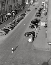 October 1940. "Grand Forks, North Dakota." 35mm nitrate negative by John Vachon for the Farm Security Administration. View full size.
It&#039;s all very different nowNone of the buildings in this photo looking NW along North 3rd Street from 1st Avenue appear to be standing today.  The Hotel Dacotah was completely destroyed by a fire that began around 11 PM on December 30, 1943.  It was said to be the most spectacular blaze in Grand Forks since the original Hotel Dacotah burned on December 17, 1897.  Fortunately, there was no loss of life or injuries as there had been in the previous Dacotah fire.
ImpressionsI'm beginning to recognize a picture by Vachon before I open the photo. He loved these high angle shots. Those are some great street lights, wonderful. There's a nice feel to this one, a bit more gritty, not as clean-looking as some of his others.  Like the pedestrian, like a Philip Marlowe character, strolling to a meeting with someone nefarious. She looks competent.
ChiricoesqueMystery and Melancholy of a Street.
That girl has a perfectshadow
Wide whitewallsNot too many that day in Grand Forks; I see only four cars sporting them out of all the assembled Detroit products. Convertibles apparently weren't a big seller in the pre-war Great Plains either.
Law &amp; OrderAlways a car parked in the wrong direction.  And where Deputy Fife to give that woman a ticket for jaywalking?
Happy DaysDid anyone actually dance at a Dine-Dance Cafe?
LookalikesI get the feeling there was only one, very busy, sign maker in town.
Stayed at the Dacotah once, sometime around 1968 or '69, when we got weathered out of our home base. The only thing I remember is a big, old style, wooden telephone booth in the lobby.
Edit: Well, maybe it was the 'Kadoka' Hotel. But I definitely stayed someplace that night!
UnmarkedI see no traffic control signs, signals or lane markings, things we take for granted today. It's a wonder there weren't accidents every day. 
N.D. license platesShould read "Land of the Long Shadow."
(The Gallery, John Vachon, Small Towns)