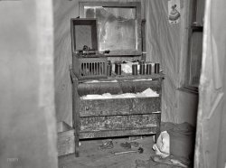 April 1937. "Bureau in the bedroom of the house occupied by the Ingrahams and the Smallwoods near Nelma, Wisconsin." A cryptic tableau if there ever was one. Medium-format nitrate negative by Russell Lee. View full size.
Way &quot;Up North&quot;As a native Wisconsinite, I can honestly say that I have never heard of Nelma, WI, which appears to be a speck on the map.
Nelma is about as far "up north" (a term used by natives to refer to the northern lakes and forest areas of the state where many have summer homes/cabins and where the deer hunting culture is king) as a Wisconsin town can be.  And, it should be pointed out, that the Ingrahams's and Smallwood's home is only "near" Nelma, which makes it even further removed from civilization. The paper on the bedroom walls gives new meaning to the term "wall paper".  (Let's hope that there is some insulation under it somewhere as the subzero temperatures in this neck of the woods can be extreme.)  The artwork on the wall appears to be Dutch, but is not Vermeer.  The bottles under the dresser could have contained liquor or liniment, both of which would have been necessary to survive life in Nelma.  I don't want to think about the possibilities of why the hammer was lying side-by-side with a pair of boy's shoes.  In addition, one can only hope that the two families were able to survive the seven years' bad luck that may have come as a result of the breaking of the mirror.  As Dave stated, this indeed is a cryptic tableau.
Old-school audioIn strictly temporal terms, playing a cylinder in 1937 would be like today sticking one of those round, silvery things - what are they called again? Oh yeah, CDs - into a machine rather than downloading or streaming an mp3 to your mobile device.
Unless I&#039;m MistakenThis tableau is one of the sets for Edison's lost 1909 version of The Shining.
Functionality of wallpaperIn response to LilyPondLane's submittal: In our modern times, wallpaper is merely a form of wall decoration. Formerly, wallpaper had a utilitarian function to seal the interior space of the house, and prevent drafts. Many houses did not use sheetrock or plaster on the walls, and cold air could seep in past the wood siding and planking.
Copyright InfringementI'd say this is a crime scene photo -- of a cylinder bootlegging operation!
Why?My question is, why is there even a photo of this scene? It may be interesting to look at 75 years later but I doubt in 1937 it was that impressive.
[It is one of the many thousands of pictures taken by Russell Lee and his colleagues documenting housing conditions for the Farm Security and Resettlement Administrations. - Dave]
This is so wrong to sayJudging by those empties and what else is scattered on that floor, I'd say someone got hammered last night. Well, I did say it was so wrong to say.
WallpaperAs an illustration for the utility of wallpaper as described by MaxCohoon, I have attached two photographs from the same series that show the house from the outside.  It is constructed of logs.  The caption for the detail photograph reads in part: "Note the earth fill around base to keep in warmth in the winter. The space between the logs is usually filled with cement or mud. The windows are removed when a house is abandoned; people cannot build glass."
Old AudioEdison actually made cylinders until 1929.
From a Victrola collectorThe mechanism looks like an Edison Amberola 30, but that cabinet is spartan.  I'm not sure what the model number is without the ornate oak cabinet.
Edison Amberola 30Agree with Michael that the machine is an Edison Amberola 30. The "-ola" suffix referred to an internal horn machine [Victor = Victrola, Columbia = Grafonola, etc.], and the cylinders made for use on the Edison machines were known as "Blue Amberols".
Regarding sstucky's comment, Edison did make cylinders until the fall of 1929, by which point they were recorded using the electrical, rather than acoustical, recording process. Although cylinders had long since fallen out of favor with the record-buying public at large, Edison continued to produce them on the basis of many rural folks still having the older machines--this photo is a good indication of that marketing strategy.
Ola!Victor claimed they chose the suffix -ola for their new internal horn machine because it had "a sound suggestive of music". Suggestive of music, perhaps, in that it was quite suggestive of another company's product: Aeolian's brand of player piano, the Pianola. Swiping other people's suffixes isn't without its poetic justice, and soon other phonograph companies began coming out with their own "olas" (actually the "o" was part of the word "piano"). Eventually an auto parts company, which had never manufactured pianos or phonographs, would appropriate it as a name for their new car radio, a name that's still around today. 
(Technology, The Gallery, Russell Lee)