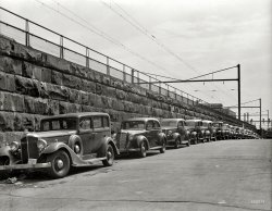 An unlabeled photo of cars parked next to train tracks from the FSA archive taken around 1939. Who can pinpoint the location? View full size.
Northeast CorridorBased on the overhead catenary over the train line I would say this is along what is now called the Northeast Corridor line between Washington and Boston.  Today the line is electrified for the entire distance, but at this time the northern end of electrification was in New Haven. 
The stone-walled embankment reminds me a lot of Connecticut, along what was then the New Haven Railroad.
Addendum to my prior commentMy guess that this was Connecticut is now reinforced by the fact that the first car has a Connecticut license plate.
I will narrow this down further and say that the photo location is looking east along Railroad Avenue in Bridgeport.  The location is most likely in a city, given the dense parking and the large factory building in the background, and Bridgeport is the only city along the New Haven line where there's a roadway paralleling tracks with the tracks being on a stone-walled embankment.  
As the twin smokestacks in the far background look too high to be factory smokestacks, I would say that they are at the power station just south of the tracks on Bridgeport Harbor.  The power station is still there, although at some point (1950's?) the smokestacks with replaced with a single, very tall striped smokestack.  
Delaware Avenue near K Street NE"I'll take 'Parking Spaces near the DC Bureau of Traffic Adjudication' for 20."
Virginia Ave SEI think this is on Virginia Ave SE in Washington, with the Capitol Power Plant in the background, prior to the construction of I-695, as we would be looking north. 
CT, west of New HavenI know the above is obvious; I am just commenting as a bookmark to come back later and see if anyone can figure it out.
Virginia Ave SW, Washington, DCI think the Connecticut license plate is a red herring.  The insulators look more like 2300 V electrification, which would be Pennsylvania Railroad. This would be the extension from DC's Union Station across the Potomac to Potomac Yards on the Virginia side of the river.
[Seacue has nailed it -- clapclapclap! Virginia Avenue SW looking southeast, just east of 7th Street. Note the matching stones. - Dave]
View Larger Map
Washington, DCAssuming that the cars are pointed north, it could be First St. NE between H and K Streets in Washington, DC.
They all look the sameSeems likely that the Pennsy folks followed fairly uniform plans in building these abutments... in most cases, they were elevating on-grade trackage to try to prevent the kind of wholesale slaughter that was routinely happening in NE Corridor cities prior to the turn of the century, as steam engines plowed through the citizenry. Having said all that... looks identical, down to the fencing and catenaries, to the NE Corridor/Pennsylvania line as it passes through downtown Newark, NJ.
Standard RR of the WorldDefinitely Pennsylvania Railroad right-of-way per the overhead catenary as well as the pipe-style railings. I'll say Newark, N.J. though I'm probably wrong. Hard to believe the PRR, once the largest corporate entity in the world, is gone now almost 40 years.
The Answer is: Washington, DCVirginia Avenue SW, as noted below by Seacue.
ID of locationThe electrical network above the tracks is far different from that of Bridgeport, CT. I think the matching of the stones is a brilliant clue!
PA SignalThe RR signals in the distance are standard signals of the Standard Railroad of the World (PRR, but I used PA in the title in honor of Father's Day this weekend). Those signals are functional simplicity at its best IMHO. Three yellow lights in vertical, horizontal or diagonal lit-pattern tell you what you need to know. Sadly, they are being replaced by color-dependent signals.
Location AsideThat Title would make a perfect title for a Sam Spade caper.   Nicely done.  Wonder what's hidden in the trunk?
Mysterymobile behind the Packard?It's a 1935 Hupmobile. Very euro-style for its day. Raymond Loewy was simply a genius, one of the greats. Shorpy should do a Loewy retrospective series -- if he hasn't already(!)
Good job, Shorpyites. Next question!How old is that stonework?
22 Cars in the PhotoApproximately. 21 facing toward the camera, and one driving away down the street. No, I will not even attempt to name them all. The aforementioned Packard looks like a 1934 model. Kudos to bohneyjames for identifying the 35 Hupmobile. The third car at the curb is a 35 Ford. The sixth car in line could be a 30 or 31 Model A Ford. If anybody can ID any of the others, it would be quite a feat!
MusclesI'm impressed by the paralell parking.  Not easy with no power steering, and the car is the size of a boat.
Definitely a 1933 PackardYou are close, but a 1934 had a lower front fender line.  This is a 1933 - no question about the year.  However, I do question the model, whether it is a Standard 8 or Super 8.  The Packard carries the V shaped headlamp and fender lamp glass of a Su8.  The Std8 had dome-shaped glass.  But, the front bumper is the size of a Std8 and not as large as the Su8 front bumper.  Wish I could read the lettering on the hubcap - it will say either Packard Eight or Packard Super Eight.
[PACKARD SUPER EIGHT. - Dave]
WWWEvery one of those cars, if displayed at a car show today, would have wide whitewalls on them, yet they were rarely used when the cars were new or near new.
As nice as the Packard is, I would choose the Raymond Loewy-designed Hupp Aerodynamic in a heartbeat. There is, maybe, one year that separates them, yet the Hupp is light-years ahead of the stodgy old Packard, stylewise. Too bad that Hupmobile was already standing next to its grave by this time. In their final days, they built a couple of beauties, this and the Skylark in 1941.
CarsI agree with the identification of the first three cars (Packard, Hupmobile, and Ford).  The fourth car looks like a 1934 Ford Fordor.  Car number five is a 1937 Dodge.  Car number six is not a Ford Model A which had the door handles on the belt line and different fenders and front bumper.  I believe this is a 1931 Chevrolet - either a 2 door coach or a 5 passenger coupe.
The Packard may have been parked there a while.  There is quite a bit of trash under the front right fender, and the birds seem to be enjoying its presence.  The bald front left tire, faded paint, and missing chrome on the front bumper also testify to a hard life. Finally, what is the marking on the driver's door window of the Packard from?  Sticker residue from a sign to tow the vehicle comes to mind, but I do not know if such signs were in use at the time.  City vehicle tax stickers existed at the time, so it might be possible larger signs could have been in use.
(The Gallery, Cars, Trucks, Buses)