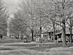 January 1939. "Main Street in Jerome, Arkansas." As well as a cozy, woodsy gas station. Medium-format nitrate negative by Russell Lee. View full size.
CompactionWith cars driving over the roots and the occasional petroleum spill, I wonder how long the gas station trees lasted.
Wait, is that George Raft emerging from the general store?
Grove&#039;s Chill TonicGrove's Tasteless Chill Tonic--a quinine mixture suspended in a supposedly-tasteless syrup.  
No SmokingProbably shouldn't be lighting up that pipe by the gas pumps.
Indiana JonesHas an evil twin who appears to be lighting up awfully close to the foreground gas pump.
Health &amp; SafetyLooks like a guy is lighting up beside the pumps. Could be wrong, but also looks like a makeshift fire drum sitting on ground behind him.
Cold Medicine of the BeastThe tree at right foreground appear (edit: appears) to have a sign for 666 cold medicine nailed to it.  The remedy, in liquid and tablet form, is still produced today by its original manufacturer, the Monticello Drug Company of Jacksonville, Florida.
[The sign was also seen here. - tterrace]
Thank you.  I now note another 666 sign on the side of the building just to the right of the tree.
Jerome GrowsJerome, Arkansas became a busy place during WWII when a Japaneese American internment camp was built which later became a camp for German POWs.  At one point during WWII it was the fifth largest city in Arkansas.  The population in 2000 was only 46 people.  The PBS documentary "Time of Fear" is an account of this camp and one located about 30 miles away at Rohwer, Arkansas.
A 1937 Plymouth is in front of the gas pumps and what looks like a 1937 Chevrolet is down the street.  Photos of similar cars are below.
All the houses appear to be raised off the ground for when the area floods.
Gas PumpsI'm curious. Did those antique pumps use underground tanks like modern pumps? Those large trees growing next to the pumps would have large root systems.
Those Tank Fill PipesI'm quite certain those capped risers you see protruding from the ground just inside the sidewalk line are for the underground tanks.
37 PlymouthWas the first car I remember my parents having, around 1960ish - age 2, along with a 56 Chevy P/U.  The Plymouth was hard to start, and Mom would often get frustrated (angry?). It apparently had very loose steering also.  I thought the faster you moved the wheel back and forth, the faster the car went.  Reality was actually just the opposite, the faster you drove, the more you had to wiggle the steering wheel to keep the car on the road.
(The Gallery, Cars, Trucks, Buses, Russell Lee, Small Towns)