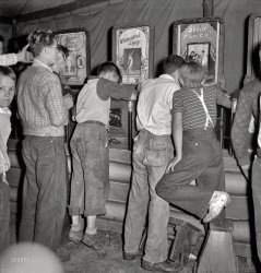 September 1938. " 'It's a dirty gyp,' say the mine workers' sons in the penny arcade at outdoor carnival. Granville, West Virginia." Stepstools helpfully provided for the shorter cineastes. Photo by Marion Post Wolcott. View full size.
After the photo was takenI bet these kids were worried about their parents seeing them watching the peep show!
MemoriesI remember seeing those at the arcades in Ocean City, MD in the 1960's. Only by that time it cost 2 cents. There was never anything really obscene. It was usually some sort of mildly naughty gag shot.
&quot;Without expectation there can be no disappointment&quot;The enticing photos and titles on the signs invariably promised more than the 1¢ show delivered.  In reality, the flip cards seldom showed more than an attractive girl fully covered by a diaphanous cloak that she almost removes by the last card.  You are always left with the impression that if there were just a few more cards, you would have received your money's worth.
Peep Shows at Rocky PointThey still had a few of these at Rocky Point in RI when I was a kid. I remember viewing one called "What Girls Do When Alone"; instead of the hoped-for steamy Sapphic interlude, the ancient, worn images showed two flappers dancing. That was it.
I guess you do get what you pay for at the Penny Arcade.
Double FeatureMy favorite filmgoers are the two guys both watching the "Strip Poker" show, trying to get a double-view for the price of a penny.
Golly gosh gee whizThis just can't be true, the youth of yesteryear were all virtuous and wholesome and never thought of, or even knew about Sex.
I Miss Those ThingsUp until about 5 years ago, they still had a few of those peep shows at the Minnesota State Fair Penny Arcade, tucked away amongst the flashy, noisy video games.  Some of them didn't work, but one or two always did, and they were still a penny.  I found them to be charming and always made it a point to watch one each time I went to the fair.  When they moved or removed the Penny Arcade, the peep shows went away too.  I tracked down and contacted the guy who owned them, hoping to buy and restore them, but he wanted much more than I could afford.  I hope they've been restored and are still entertaining people somewhere.
Truth in advertisingPlayland-Not-At-the-Beach in El Cerrito has a few of these old peep shows.  One, called "How to Clean Up at the Races," is a tiny diorama of plastic horses, a shovel and manure.  Another, "Fan Dancer," is exactly that - a shaking paper fan with doll legs.  You got exactly what was advertised with these things, so don't call them "a dirty gyp," kid.
We expected to finally see the censoredIn  1957 at Bertrand Island  Lake Hopatcong NJ we kids would drop our pennies in and see "The electric chair at Sing Sing" a prisoner being executed. and a Victorian era  Fan  dancer...hot stuff. The flip card viewer was a real attraction. 
(The Gallery, Kids, M.P. Wolcott, Movies)