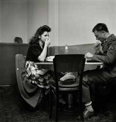 May 1942. "A corporal in the Army takes his girl to dinner. Bakersfield, California." Photo by Russell Lee, Office of War Information. View full size.
If he&#039;s luckyMaybe she won't notice his goofy socks.
This photo speaks volumes1) You have got to love the rolled socks!
2) Take a good look at the young woman's look of love toward her soldier - priceless!
3) It goes to show you that Heinz Catsup was and is an American standard.
A study in contrastsWonderful shoes, those brogues, but horrible socks.  Also, while she’s eyeballing him, he’s intent on forking that food down.
The Inevitable Ketchup BottleMy dad served in the Army from 1941-1945, and as a child I remember that he put ketchup on everything he ate, including mashed potatoes and scrambled eggs. I asked him why and he said that during his time in the service, the food, especially when he was in the Pacific, tasted so bland that ketchup was put on every meal to enhance it's flavor. He did this his entire life, and despite the fact that mom's cooking was so darn good, my brother and I put ketchup on our food too. Mom yelled; dad simply smiled.
Civvie FootwearEither this corporal is a reservist who's not yet received his complete clothing issue or his squad leader is blind, because the shoes appear to be wing tips, decidedly non-regulation.
Movie StarThe young lady is movie star attractive.  Army shoes back then may have been brown rather than black.  The rolled socks are a hoot.
War is HellIf he was sent overseas (and I'm sure he was) I can imagine how tough it was to leave that beauty alone among the 4F wolves.
Brownshoe ArmyUntil sometime in the early 1960s, dress shoes were brown to match the olive green Class A uniforms.  The present forest green uniform came in to replace the prior uniform and necessitated black dress shoes.  During my time as a draftee the phrase "brown shoe Army" was used to denote something as our of style.
Although wing tips were not likely standard issue it is entirely possible that in May 1942, some civilian footwear was issued as a substitute until uniform production could catch up with man power.  
Those socks, though, that just indicates a sorry soldier.
Maybe he is a memberof the 117th Beau Brummel artillery unit.
Ah, You Kids...never had to deal with socks where the elastic - which was only at the top, anyway - gave out after a week and the whole sock slid uncomfortably down inside your shoe, leaving your heel naked and blistered. That rolled top is one solution.
Re: Brownshoe ArmyRegarding Texcritic's comment, brown shoes began to give way to black when the dress green Class A uniform was adopted in 1955. It took a while to get all those brown shoes out of the system, and to get the new black ones in. 
I've talked to several Army veterans who served at this time, and they all remembered being issued two pairs of brown combat boots, two pairs of brown dress shoes--and two bottles of black shoe dye.
The old WW2 style uniform with Ike jacket was slowly phased out during this time. I had an uncle who served from 1957 to 1960--in basic he wore the old style uniform, but had the dress greens by the time he got out. The old style uniform was officially declared obsolete in 1960.
Army vets of the eraI've talked to Army veterans of this era and although I've never seen it before personally, I've been told that wearing white socks with the uniform while on leave was considered to be "cool."  Of course, wearing one's socks rolled was also considered to be "cool, as well.
Even when I was in the Marine Corps during the Vietnam era, it was common to see soldiers in the airports wearing non-standard uniform items in their comings and goings around the country and overseas.
These practices were also common among sailors, who would pick up non-standard uniform items in the ports where there were thriving industries catering to this market.
I definitely wouldn't go so far as to call this soldier a "sorry soldier."  We certainly know nothing of him or what became of him. He may be a decorated combat veteran for all we know.
I would guess that the soldier was on leave, because if he were to be on liberty, I'm sure the MPs would be on the look out for such things.
Looking military...didn't mean what it does today. There was a jaunty aspect to WW2 servicemen, with their tilted covers and brilliantined hair, that would be totally unacceptable today. 
Her ShoesFrom what I can see of her shoes they look very much like a pair my mother was wearing in a picture I have, taken at roughly the same time.  All the pictures I have of her and my aunts in that time period look so elegant.  As with this lovely lady, hair done just so, skirt and top nicely matched.  And lets not forget the shoes!
Look at his Army hat.The hat seems to be a very high-quality hat, not an issue item.  I wonder if the corporal wasn't a California National Guardsman or Army Reservist from the Bakersfield area now on active duty.  National Guardsmen/Reservists would purchase commercial uniform items instead of or to replace issue items.
Unit PatchAfter noticing his shoes I saw that he wears no unit patch on his shoulder.
Changing of the colorsI believe it was 1957 when the U.S. Army changed the boots and shoes from brown to black. New ones weren't issued, but we had to dye the existing footwear. We could have done them ourselves, but most of us paid to have the color changed.
The shoesThose shoes are NOT wingtips, but they do have broguing. I recall seeing a picture of an American general wearing similar brown brogues.  So they were definitely acceptable with that uniform.  And I suspect the socks are a light tan. I think that was the uniform sock color at the time. And yeah, before Spandex, socks fell. Period.
And while I'm here, may I just say that this soldier's girl is a DOLL. Pretty woman!
(The Gallery, Eateries & Bars, Russell Lee)