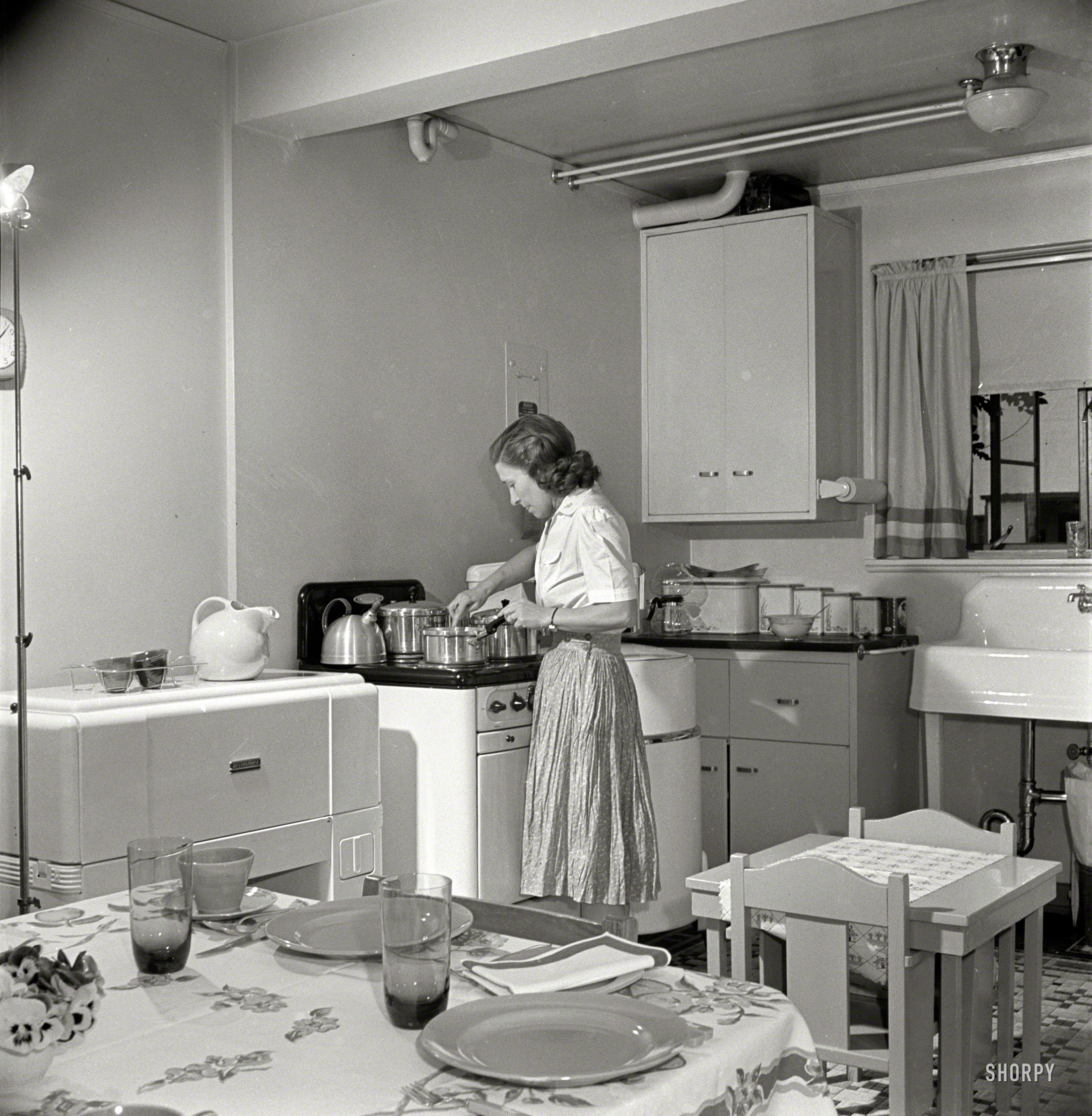 May 1942. "Greenbelt, Maryland, federal housing project. Mrs. Leslie Atkins preparing dinner in her kitchen, one end of which is the dining room. Notice the mangle and washing machine on either side of the stove." Medium format negative by Marjory Collins for the Office of War Information. View full size.