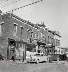 August 1942. "Street in Dillon, Montana, trading center for a prosperous cattle and sheep country." Evidently a parched and thirsty territory, slaked by what we'll christen the Unangst Block. Nitrate negative by Russell Lee. View full size.
Old BuildingsThank goodness the people of this town have not torn down the old buildings. They must be fortunate in that there is still a need for those buildings and that they are not sitting empty and going to ruin like buildings in so many smaller towns. 
Hang That Beer SignA curious follow on to the previous image.  We get to see how the hooks on the beer sign, avail themselves.
FM
Radio, rooms, and beerNobody has commented but that looks like a long distance radio antenna on top of the Unangst building, Would love to know more about that!
Dive BomberThat second car (the one with the driver at the wheel) looks like it practiced a time-honored practice we used to call "dive bombing" whereby the car is parked nose-first against the curb whilst the rear end protruded into the street.    
Is This Trip necessary?A fellow could attend a lodge meeting, get a haircut, get thrown out of several bars, and find a flop, all without burning any gasoline or wasting any rubber.  Splendid urban planning, and very patriotic.
A close shave*That* is one handsome grille on the Nash in front.  It's like a gigantic, gasoline-powered electric razor on wheels.  Also, the hats on those two fellers next to it makes me think they're likely sheepherders and not cowpunchers.
Give the fella a handbecause he needs one trying to parallel park that second car.
Next to the MINT signWhat is that rectangular object at the lower right of the MINT sign?
Buffet Bar?Is that all you can drink for one low price? 
Nice pic on Google Panaramiohttp://www.panoramio.com/photo/12714350
Sing &quot;Melancholy Baby&quot;From the disheveled appearance of the man exiting the bar, the gingerly manner in which he is walking and his tottering posture, I'm thinking he may have had one too many.  Not to worry though, the guy in the second car may be driving him home.
It&#039;s all still thereLooks as if some of those buildings still house bars, including the Klondike Inn at 33 East Bannack Street (the Mint was there in 1942).
Odd FellowsThe building at the far end of the block seems to be the meeting hall of the IOOF - the Independent Order of Odd Fellows. I guess that makes the two guys on the left just ordinary fellows.  
&#039;40 NashFirst car is a '40 Nash (the '39 did not have sealed beams), the one nosing into the curb looks like a '37 Ford Standard.
Love that 1940 NashMade in Kenosha, WI at the Nash-Kelvinator plant.  They were at the vanguard of climate control interiors in thier day.  
Unfortunately the Kenosha plant which was owned by Jeep did not survive the 2008 great depression.
How StrangeI remember Nashes from the late '40s on. How strange, to see a Nash that's handsome instead of peculiar!
(The Gallery, Cars, Trucks, Buses, Eateries & Bars, Russell Lee)