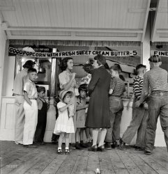 August 1942. "Butte, Montana. Children are transported to Columbia Gardens, an outdoor amusement resort, every Thursday during the summer by city buses." Medium-format nitrate negative by Russell Lee. View full size.
Better than goodThe boys on the right with the bad haircuts seem to be investigating what's for sale in the next booth.  According to its sign, it's better than Good, it's Fine!
That buckle can't be worn anywhere else but on the back of his dungarees, because it's not on a separate belt, but on a denim cinch that's sewn on as part of the jeans.
Chino BucklesWhen I was in high school in the 50's, the chino buckle served a purpose for guys.  If it was unbuckled, you were available for dating the ladies.  Buckled, you were going steady with someone.
Letter Until You Run Out of RoomGot enough adjectives on that butter, have ya'?
Butter adjectivesActually, "sweet cream" is a term of art in butter making. It indicates that the butter was made from uncultured cream as opposed to cultured cream. The latter is a generally richer flavor and predominantly European, though available in the more civilized parts of the US.
Women in pantsInterestingly two of the women in the picture seem to be wearing pants. That must have been something of a novelty.
[Definitely not in the rural West. - tterrace]
Brave parents!Trusting the two very dapper children wearing crisp, summery white clothes to a dusty amusement park in August.
Popcorn with sweet cream butterWell after reading JWP's interesting explanation about the butter, I could murder a few bags of that popcorn!  Yum!  Don't you just know that it had to be absolutely delicious?
Those Back-Belted PantsThat late 50's/early 60's style for trousers (can't remember buckles on jeans) were called "Ivy League" style. Ivy League caps (think sports car drivers' caps) had little "belts" on the back too.  These are also associated in my mind with "Dobie Gillis" three-quarter length shirt sleeves from the same general era.
[I had a pair of pants like that in high school, 1960-4. I was never quite sure if I looked cool or silly. - tterrace]
Fashion PoliceThese belts on the back of the pants were popular when I was in grade school in the middle to late 50s. However the nuns weren't too crazy about them because they would scratch the wood on the back of the desk chairs. 
Bucking the trendNot everyone in the picture was getting popcorn; the small boy on the left appears to have an ice cream on a stick.
Tonsorial ArtsThe two boys on the far right seem to be victims of the same barber, probably a close relative.
A Real CinchThe kid on the far right shows an early sense of style, wearing dungarees (my wife hates it when I use that dated word) with a buckle in the back. I had always thought this was a fashion statement from the late '50s when it became the in fashion for me and most others to wear chinos with that same useless buckle. Along with the obligatory madras shirt, of course.
The kid in the middleLooks like he is wearing his Keds.
Butter, Womens Pants, Madras and bucklesYes indeed, cultured butter is the best. I make my own from time to time adding cultures to "sweet cream" for a far more flavorful product. Re the panted females: I have a picture of my aunt wearing pants in 1943- the year of her death at age 18. She did not live out West but in southern Illinois. The back-buckled jeans was a surprise to me as well; I thought this was a late '50's thing when I was in grade school, along with madras shirts. Forgot how fashionable we all were back then. A final comment: I was the victim of a tonsorial artist who was a close relative until we got in an argument about her lack of skill. She quit right in the middle of a contested haircut and told me I could just go to the barbershop - and pay for it out of my pitiful allowance. I was broke but happier after that. We are now speaking again, but she still bosses me around. Mom, cut me some slack - I'm 64  
Columbia Gardens historyColumbia Gardens was a fantastic place in its day. My mother and her first husband used to go dancing there before the war. I remember riding the roller coaster during high school. An article from The Montana Standard is quoted here.
(The Gallery, Kids, Russell Lee)