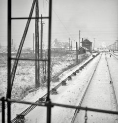 January 1943. "Freight operations on the Indiana Harbor Belt railroad between Chicago and Hammond, Indiana. The train passes many interlocking towers on the way and the conductor watches them for any special messages." Medium-format negative by Jack Delano, Office of War Information. View full size.
Wooden DodoVirtually every track crossing of any importance had an interlocking tower guarding it; some were single story shantys, and some were massive brick or block structures. But most were very much like this one. As busy as this tower looks capable of, it likely was manned 24/7. 
The rods visible left of the track physically align switches, derails and even semaphore signals. More modern interlockings had electric motors to move these things, although I worked around such a plant as this well into the 1980's. 
Towers such as this are Dodo birds, or very nearly so. The function of protecting a level crossing,interlocking or a junction is today controlled from many miles away, and the towers have been falling all over the country for decades; if any are still in use for intended purpose, they are few in number.
InterestingI don't know why, but I love the wintry-scene railway photographs.
I am curious what the lines running along the track are, on the left.  Are they signal lines?  They seem too far away from the tracks to be an electric engine power source, and the supports would interfere with power engine power shoes.
>>>>>>> Thanks to Olde Buck!  They are control rods for signals and turnouts, which makes much more sense.  I've read that those controls took some muscle to use, and no wonder!
An Armstrong OperationWhen I was about 14 a tower man in Cumberland, MD let me try my hand at an interlocking lever. It wouldn't budge. The name is apt. That tower also had a timer which had to be set before any levers could be thrown, then each lever was locked in its new position until the timer expired after the next train passed. It was to make the operator think out his moves carefully, and prevent last minute changes.
I H B R RSeems the name went from  Indiana Harbor Belt Rail Road to Indiana Harbor Belt R R, to Indiana Harbor Belt, then to Harbor Belt finally on its later diesel engines it was just Harbor.
Location on &quot;The Harbor.&quot;Looking to the northwest at McCook, the Santa Fe crossing. I recognize the jog in the mainline on the other side of the diamond. 
Manual interlockings The Harbor (IHB) had about a dozen or so interlocking towers back in the day, each one a busy place. I’m going to hazard a guess and suggest, for several reasons, the featured picture is the old IHB Ivanhoe tower, controlling movements of IHB and EJE traffic across the diamond about a mile east of Gibson at 160th Street. To the best of my knowledge: 1) Dolton Tower, controlling movements across the UP (north/south) and IHB (east/west) is still manned by an interlocking operator, 2) the GTW tower at Blue Island is still a manual interlocking, as is 3) Gibson Yard west end tower. At one point most, if not all, of these towers were "strongarm" plants. Remote controlled switches and signals have replaced the Stone Age technology, but the operator headaches can still be intense. He/she has to balance the interests and wishes of a yardmaster, a dispatcher, and a trainmaster, among others, each of which has their own priorities. Anyone who has ever worked a Chicago tower will know well what I speak of.
Attached is a picture of a strongarm operator lining a switch, one perhaps several hundred feet from the tower.  
McCook: Home of EMDJust on the other side of that aggregate pile in the background is General Motor's Electro-Motive Division, at 55th St. and old U.S. 66. All new EMD diesel locomotives delivered from this plant were handled by the IHB first. 
Manual interlockings photokreriver, what tower is shown in the photo you posted? It doesn't look like an IHB tower to me.
(The Gallery, Jack Delano, Railroads)