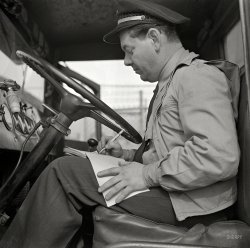March 1943. Washington, D.C. "Bob Daugherty filling out his log book after lunch. The Interstate Commerce Commission requires all truck drivers to keep time records." Photo by John Vachon, Office of War Information. View full size.
Trailer Brake LeverModern tractors still have the separate trailer brake lever, but it's usually on the dashboard somewhere, not on the steering column as pictured here. It really doesn't serve any purpose!  
Pull your JohnsonThe trailer brake valve is called a Johnson bar or johnny. The term might come from railroading, where a 'Johnson rod' was a mythical part greenhorns were sent to find, or was a catch-all phrase for unexplainable problems with the engine.
 Until Spring brakes became mandatory in the '70's, trailer parking brakes depended on having air in the trailer tanks. When the air leaked out over time, a driver trying to hook a trailer could end up chasing it all over the yard as it would roll away on contact with the tractor. He would thus back up to the trailer, hook up the airlines (with 'gladhands', another term borrowed from railroaders),charge the trailer tanks, pull his johnny to lock trailer brakes, and hook her up.  They are still required on tractors, although seldom used.  Using it to get out of a jacknife would only worsen things, like using the handbrake in your car during a snowfall.
Bob&#039;s brother, Ralph.Bob had a younger, look-a-like half-brother, named Ralph who also worked as a driver. The last time they met, Ralph was on his honeymoon.
I am trying to picturea modern teamster wearing a tie to work.
Funny book?The truck drivers name for the log book is 'funny book'. It probably got that name from the fake entries made in it to conform with the letter of the law.
Book of liesAnother term for the logbooks was "Book of Lies".
Natty AttireThe gizmo just to the left of the steering wheel is the handle for the spotlight.
They disappeared long ago, along with uniformed truck drivers.
SpotlightThat spotlight is an Appleton, long gone from the stores but a highly coveted find now.
Spotlights (now almost all the generally similar Unity) remain common; you will see at least one on every police car and emergency vehicle out there, and on many taxis and big trucks.  Civilian spotlight geeks remain out here, too! I note from my own post that we are at least as obsessed with bizarre obsolescent details as the train geeks.
That lever on the steering columnThe lever on the steering column is clearly not a shifter nor a turn signal. It has a little pipe coming out of it.
I'd guess it's for a trailer brake or a compression brake, but someone with more practice driving 1940s big rigs would know better. 
Make, year and model of the tractor?I'm thinking it's a late 30s or early 40's Autocar, before the comfort cab.
Trailer Brake LeverThat trailer brake lever is very useful. It can be used to apply only the trailer brakes if the tractor starts to jackknife. Applying the trailer brakes helps to straighten out the rig whereas if you use the brake pedal to apply all brakes you might increase the chance of a jackknife.
(The Gallery, Cars, Trucks, Buses, D.C., John Vachon)