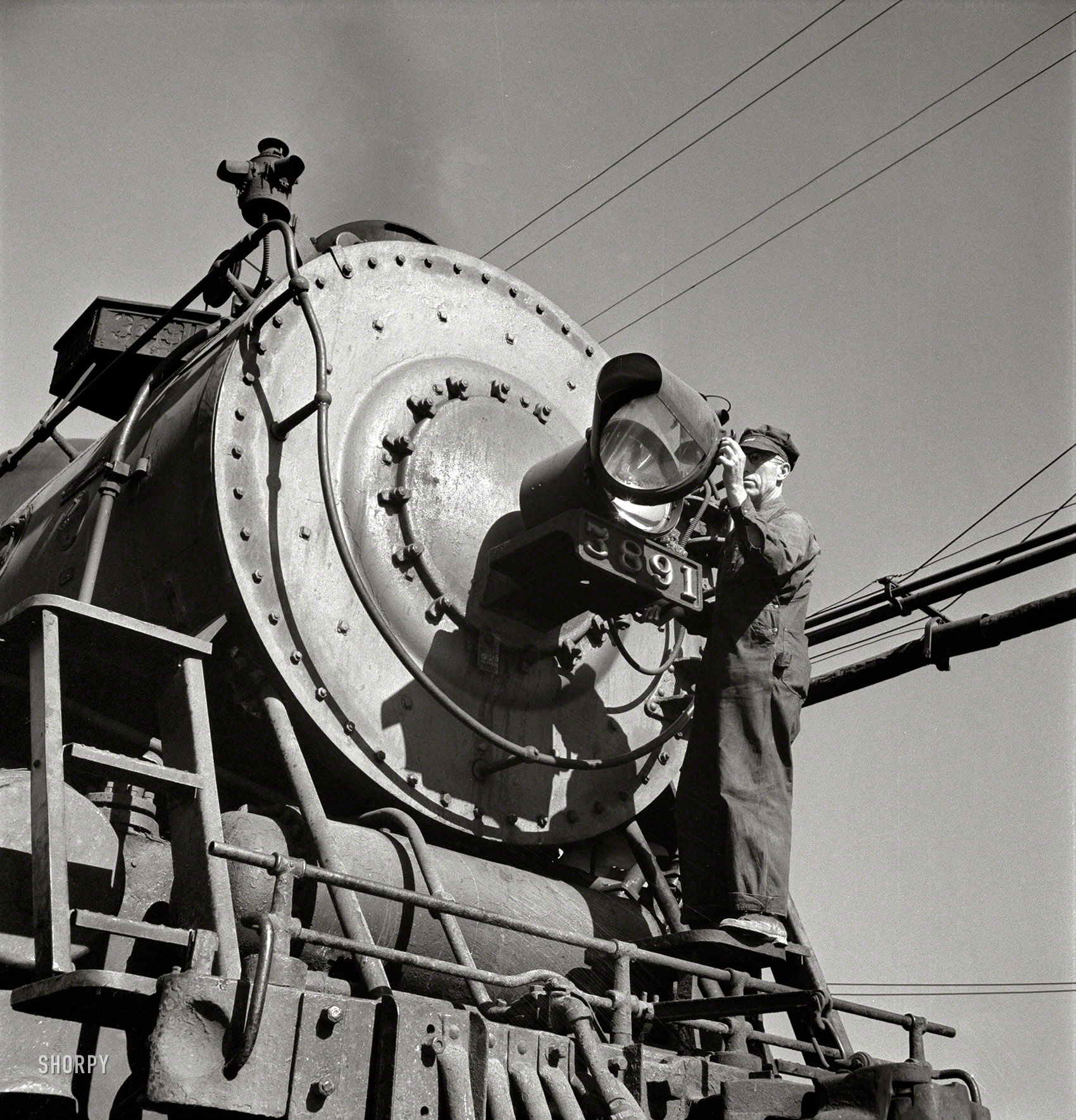 March 1943. "Needles, Calif. Electrician B. Fitzgerald cleaning the headlight of a locomotive at the Atchison, Topeka & Santa Fe yard. All engines operating west of Needles are equipped with hooded headlights in accordance with the blackout regulations." Photo by Jack Delano, Office of War Information. View full size.