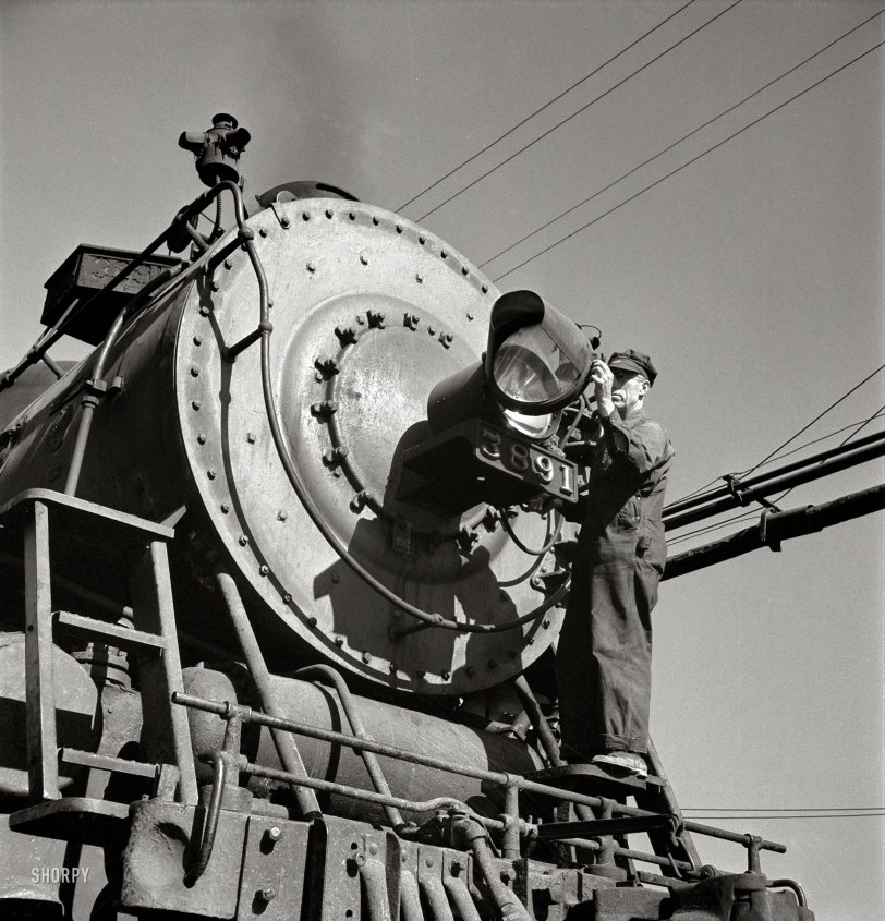 March 1943. "Needles, Calif. Electrician B. Fitzgerald cleaning the headlight of a locomotive at the Atchison, Topeka &amp; Santa Fe yard. All engines operating west of Needles are equipped with hooded headlights in accordance with the blackout regulations." Photo by Jack Delano, Office of War Information. View full size.

