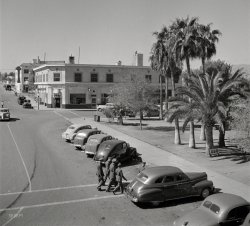 March 1943. Needles, California. "General view of street leading to depot of the Atchison, Topeka &amp; Santa Fe Railroad." Photo by Jack Delano. View full size.
Cooling towersThe structures atop both hotels resembling inverse pagodas are cooling towers, as no decent hotel in a place like Needles could have done any business without some kind of comfort cooling. They may be water-cooled condensers for refrigerated systems, or they may be just plain old "swamp coolers"; it's impossible to tell from this evidence alone. From what I've seen and been told, the louvers on the sides were usually made of wood, probably cedar or cypress. The man straddling the foreground tower attests to the intensive maintenance that these systems required, while the tower in the background may have emanated the warm smell of colitas.
The Joads, of course, drove past this place at night, after spending the searing hot desert afternoon immersed in the Colorado River, the poor man's air conditioning. 
Welcome to the
Less ornate today.I am certain this is the location, with all the charm thoroughly removed:
View Larger Map
ID for CarsWe often hear it said that today's cars are not distinctive enough to ID. That was also true in the 30's, especially from the rear!
To the right of the fellas on their way to the USO Club is a nice new 1941 Pontiac Streamliner "Torpedo" Four-Door Sedan. With a nice pair of non-factory fender skirts as well.
To their left is a 1939 Ford Tudor. Can't tell if it is a Deluxe as we cannot see the right tail light - or lack thereof.
Updated IDThe 1939 Ford is definitely a Tudor and not a De Luxe Tudor.  The De Luxe models are easy to spot because the headlights are in the fenders, not between the fender and hood as shown here.
The 1941 Pontiac is a De Luxe "Torpedo" Four Door Sedan and not a Streamliner.  In addition to there being a definite break between the roof line and the trunk, the side trim on the De Luxe ends in a straight line, parallel to the ground, before the trunk begins.  The Streamliner, which is a true fastback, has side trim which extends far beyond the trunk top, dips down towards the rear bumper, and follows the fender line.  
Also of note, the fender skirt shown on the Pontiac is a standard accessory that was shown in the Pontiac catalog.  Photos of everything below.
Needles Today...The soldiers in this picture were most likely stationed at the Needles Army post located between the Riverview Cemetery and what is now Needles Airport on the road going to Blythe. They would be going to the U.S.O. located inside the El Garces Hotel (Harvey House), which also serves as the passenger train station (it was completely refurbished in 2014 and serves as an Amtrak station and office space, although the only office space being used now is for the local city bus company).
Most of the buildings in the picture are still around. Some are abandoned but available for reuse. We are looking south on F Street from Front Street, which jogs around Santa Fe Park via F and G Streets. Looking at the postcard here, the building on the left, at the corner of F and Front Streets is the Needles Point Pharmacy along with some other businesses. Beyond that (during wartime) there was a service station at the northeast corner of Broadway and F Street.
Across the street from that is the Needles Theater (Cinema)/Masonic Lodge (the local Masons built it in 1929 as a dual purpose building). The theater closed down many years ago and the Masons no longer meet in Needles. However, the marquee still works and it can be rented for messaging. On the other side of F Street, where the palm trees are is Santa Fe Park, which is in front of El Garces Hotel (Harvey House). There aren't as many palm trees today.
We cross Front Street as it jogs around Santa Fe Park. Here we see the Needles Point Liquor Store (complete with loyal customers at the front door) and then we see the Needles Eye Lunch. This is the Butler Building and it's all abandoned but it's serviceable. Beyond the Needles Eye Lunch we see a Shell Station and the northwest corner of F Street and Broadway. This is now where Needles Glass and Mirror do business. Across the street from that we see the Hotel California, which burned down in 1952. Today that site serves as the parking lot for the Frontier Phone Company. 
Beyond that is Robinson's Motor Inn (cottages) which is now Robinson's Apartments. (Three of the old 1930s motels became apartment complexes.) Needles Point Drug and Liquor Stores are now neighbors on Broadway to the southwest of this picture. The former site of the Needles Point Drug Store is now property of the Needles Unified School District. There are still other businesses in that building, including a beauty salon. The solid white line down the middle of the street is now a double yellow line. The crosswalk from the drug store to the park no longer exists. Only parallel parking is allowed on Needles streets today.
(The Gallery, Cars, Trucks, Buses, Eateries & Bars, Jack Delano, Railroads)