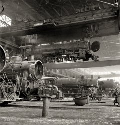 March 1943. Albuquerque, New Mexico. "An engine being carried to another part of the Atchison, Topeka &amp; Santa Fe railroad shops to be wheeled." Photo by Jack Delano for the Office of War Information. View full size.
Helmet=Steam Dome CoverAs you can see on the 1823 the sand "dome" or more correctly sand box, is still attached to the boiler (it is seldom ever removed) the diagonal piping being the discharge lines for forward or reverse sanding.
If you look at the steam dome, the projection which is the highest point at the middle of the boiler, you can see that the sheet metal cover is removed, and laying on the shop floor.
Boilermakers had to enter the boiler through an opening at the top of the steam dome for checking flues, throttle linkage and the dry pipe.
upside down helmet...Further on the upside down helmet: The sand dome would be larger with two cutouts for the sanders for both sides of the engine. The cutout seen on the left is more than likely to accomodate the pipe out to the whistle.
Browsing the Denver Public Library online AT&amp;SF images are a great way to see this feature on Santa Fe engines.
The HelmetThe "helmet" is the cover off of a sand dome. The hole for the sander pipes can be seen in the rim of the cover there to the left.
AsbestosThe man working on the steam induction pipe on engine 1696 appears to be insulating the pipe with asbestos putty. You can also see some of the Asbestos lagging around the front of the same engine. When old steam engines are renovated for museum pieces today Asbestos abatement is one of the first things done.
Shopping BadThe buildings in this complex have been seen in several "Breaking Bad" episodes.
250 tonsOf lifting power.  Very impressive.
1696 in actionThere's a good shot of engine 1696 running in 1948 on this site.
The Army HelmetWhat is with the freakishly large army helmet on the floor?
Marty and DocRailroad tracks?  Where we're going, we don't need railroad tracks.
The HelmetI'm usually one of the people supplying information regarding railroad photos, but now I have to beg a question: What is that object sitting on the floor that looks like a giant steel military helmet?  I've seen this pic before in much smaller size, and this is a detail I never noticed.  Any ideas?
Steam locosIf you notice, each of these three locomotives is a different model. One of the problems with steam trains was that each railroad had just a few of any given design of engine, and so had to carry parts, blueprints, procedures, etc., for the maintenance of many different machines.
The diesels that replaced them were not really any more energy efficient or powerful. In fact many steam locomotives actually gave more traction for energy expended than the diesels. The diesels' great advantage was standardization, and needing less maintenance.
So while the steam locos were magnificent creations, their days were numbered, even as this picture was being taken.
Where&#039;s Waldo?At first you don't notice them, and then -- I count at least twelve workers in this fascinating view. From left:
Man on ladder; blurry man inside smokebox; the distant crane operator; face inside near vise jaw; blur walking toward front of 1823 by last driving wheel; man working on valve gear of 1823 (head &amp; shoulders above vise); man standing on pilot behind open smoke box door of 1823; group of five, distant right.
Did I miss anyone? 
GroundedI believe this is the same flying train, this time with wheels on and on the track.
Steam dome cover.Sandboxes don't have covers. You can see on 1696,  the nearest loco,  that the sandbox is a substantial component made up of castings and rolled plate of at least 3/16th of an inch thickness. On more modern locos the entire unit was fabricated by welding. 
The "helmet" is in fact a steam dome cover - notice that on 1823 the cover is removed exposing the actual steam dome on the second course of the boiler.
The hole in the side referred to by DBurden is where the whistle is mounted on an elbow that in turn is mounted to the steam dome - as can be seen on the suspended 3733.
Air conditioning, againI wonder what it was like in that shop in the height of summer, and the low of winter. 
Albuquerque ....Yup, old shop buildings are still there in Albuquerque. Things are in slow-motion to turn them into the Wheels Museum with a home for the 2926 steam locomotive that is being restored right now !
GrandpaMy grandfather was working here at this time.
Magnesia, not asbestosMEGGAHURTZ: The lagging on the steam pipe is made of "85% magnesia" blocks, not asbestos putty. The material was 85% carbonate of magnesia and 15% asbestos fibre as filler.
Jazznocracy: Contrary to your claim, it was not unusual for US railroads to have hundreds of the same type of locos in a given class. Santa Fe had 352 2-10-2s alone. Moreover, their backshops were designed and equipped to build the parts needed to maintain these locos in-house. Drawings were either provided by the loco builders at the time of manufacture, or the railroad's own mechanical branch. 
Diesel parts, however, had to be bought in from the original manufacturer. The diesels running in 1943 were not particularly standardised, nor were they light on maintenance.
Mountain and Prairie and Santa Fe1696 is a 2-10-2, built by Baldwin in 1912 or 1913.  It is a "Santa Fe" type, so named because the railroad requested a modification to a 2-10-0 locomotive.  This type was pretty popular; over 2200 were built for various railroads between 1903 and 1931. It was scrapped in 1954.
1823 is a 2-6-2 "Prairie" type, built by Baldwin in 1903. 1823 was scrapped in 1949.
For the details on 3733, see this Shorpy photo.
Mass employmentConsider this is just one shop of the Santa Fe, and there were many others.  Each shop employed a thousand men (or more), and consider the hundreds of independent railroad companies in the wartime nation.
Each railroad had steam locomotives specifically designed to the geography in which they operated, and they appeared in wheel arrangements from four wheeled switchers to massive, hinges chassis to snake around curves when the boilers were too long!  That was railroading at its grandest period.
(The Gallery, Jack Delano, Railroads)