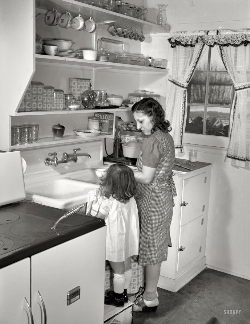 January 1942. "Bantam, Connecticut. Defense homes. Little Ann Heath is eager to try out all the facilities of her parents' new four-room defense housing unit, after spending most of her life in a single furnished room. Here she pushes her footstool to the sink in order to help her mother clean up the dinner dishes. Mrs. Heath, a native of Winsted, a city some 25 miles away, is delighted with her new kitchen -- the first she's ever had which she actually considers as a kitchen, and is trying out all the recipes she has collected in five years of married life. The Heaths pay $30 monthly for their apartment." Medium format nitrate negative by Howard Hollem for the Office of War Information. View full size.
