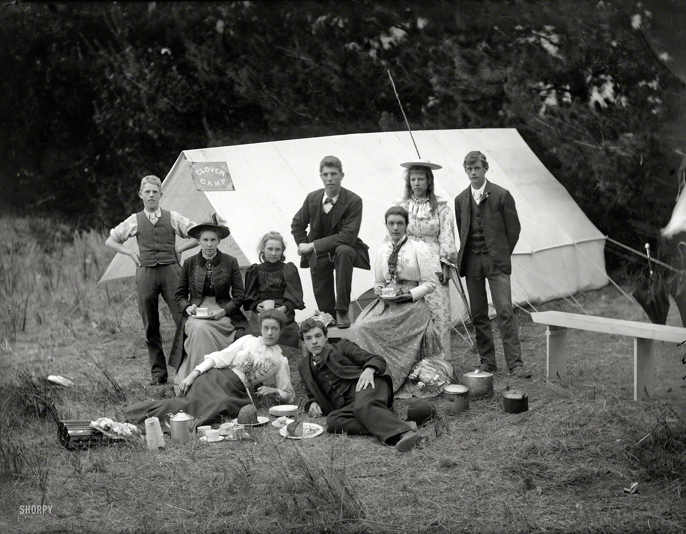 Circa 1905 near Christchurch, New Zealand. "Young people with camping gear, having tea and cake in front of tent with 'Clover Camp' sign." These natty campers have even brought umbrellas. Glass plate by Adam Maclay. View full size.