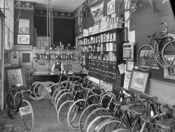 1910. "Cycle shop interior. Christchurch, New Zealand." B.S.A. stood for Birmingham Small Arms. Photo by Steffano Francis Webb. View full size.
BSWAgree with Papa Bear, Brit bicycles, motorcycles and cars used a bolting system called "British Standard Whitworth".  It differs from the US system in several ways, BSW wrenches are labeled for the size of the bolt instead of the US system of labeling the size of the the nut hex.  And common bolt sizes 1/4" to 1/2" used the same 26 threads per inch, instead of the US system of varying the TPI with bolt size.  The angle of the threads was unique as well, 55 deg. instead of 60 deg. (from memory).
In other words, almost nothing standard in the US fit Limey bikes!
The Brits eventually sold out to a metric way of life, thankfully!  
EverywhereA National Cash Register from here in Ohio, even in New Zealand.
EquipmentI had no idea frame pumps were around so early.
I like the two bikes in the foreground that have the skirt nets. I'm happy that in the Twin Cities of MN area these are once again on bikes because more people are riding non-athletically, dressed to go out.
No chain guardsWhich makes it very hazardous to ride if wearing long pants since they often got caught between the chain and sprocket and when this happened your leg was pulled down as the sprocket turned. The usual result was falling off the bike. I hope they were sold separately.
Other BSA productsBSA also made motorcycles, and as the name implies, was a major producer of the No.1 Mk III Enfield rifle, standard British Army rifle of World War 1.
Wheels of EmpireThank you for publishing these New Zealand images. This one is particularly fascinating. As a boy in 1970s Britain I rode a B.S.A. bicycle myself. The shop is also advertising Eadie brand bicycles, taken over by B.S.A. in 1907. These had originally been made by Royal Enfield: 'built like a gun, goes like a bullet'. The brand names are wonderful: Britannia Tyres, Dreadnought Tyres ... tyres were clearly a seriously patriotic business in the British Empire c.1910.
Clip your pants legPants leg clips were used to keep the pants leg out of the chain. These are still available, and cheap; Google "bicycle pants leg clips". 
BSA Lightning RocketI rode a 1965 BSA Lightning Rocket and BSA meant BSW wrenches for their fittings
Interesting brake system.It appears the brake pads pull upward against the inside of the rim, somewhat like a drum brake, rather than squeezing it disc brake style like modern bikes do. 
Bluemel partsI took notice of the little card hanging on the wall just above and left of the cash register, holding what looks to be several horizontal pencils.  The top says BLUEMEL with the "E" appearing to be a different color.  Vintage bicycle experts are probably familiar with the brand name Bluemel Bros.  Did a little searching and found it was a company in England that made bicycle and motorcycle accessories.  One of the Bluemels came to the USA in 1906 and became a founding father of speech pathology on stuttering and stammering.
Found this website on the bicycle part of the family, started in 1860.
WhyWould they offer to 'buy back your National Cash Register checks'?  For money or merchandise?
Brakes were a rarityA few of these bikes have front caliper brakes; none of them have coaster brakes. They all appear to be fixed gear bikes. Curiously, this 100-year-old-style is all the rage these days. 
Cars too!BSA also made cars. 
I WishI could get "DREADNOUGHT TYRES" for my 4WD truck. With big white letters on the side.
Carbide LampsThere is a nice selection of carbide lamps (and bicycle bells) on the stand on top of the right side of the counter, and kerosene lamps on the floor stand to the left. Calcium carbide was placed in a lower chamber of the lamp, and above it was a reservoir of water. When a valve was opened it dripped water on the carbide, which produced acetylene gas. Some of these lights may also have operated on kerosene or gas. You can see other examples here.
Rod brakesVintagetvs is correct that all of the bikes here with front brakes use "rod brakes". When the brake lever is squeezed a rod and pivot system, instead of a Bowden cable, lifts the brake pads upward against the inside of the rim. These were once very common and are still widely used in various (mostly less developed) parts of the world. They are relatively rugged, and easy to maintain and repair with simple tools.
The coaster brake was only about 10 years old at the time of this picture. I don't know how long it took for the coaster brake to become commonly used, but since it would be very much more expensive to produce I suspect it remained an expensive option for quite a while. Epicyclic hub gear systems are a little older than coaster brakes, and truly functional derailleur systems are about 25 years later than this photo.
BrooksLooks like lots of Brooks leather saddles on the bikes. Still available. I have one on my road bike. Kinda hard on the tush until they get a little broken in, but then they are the best!
Bike tubeThe bike on the left, and a few others have a tube sticking out from under the seat, facing forward. There is another tube facing back from the handlebars. Anyone know what they were for?
Seat and handlebar tubesmailman7etc. It would appear that the forward-facing tube under the seat is the mount for the saddle. The rear-facing tube under the handlebar allowed the handlebar to be slid closer or further away, held in place by a clamp.
British Standard WhitworthPapa Bear's mention of BSW wrenches reminded me of this piece on Joseph Whitworth.
(The Gallery, Bicycles, New Zealand, S.F. Webb, Stores & Markets)