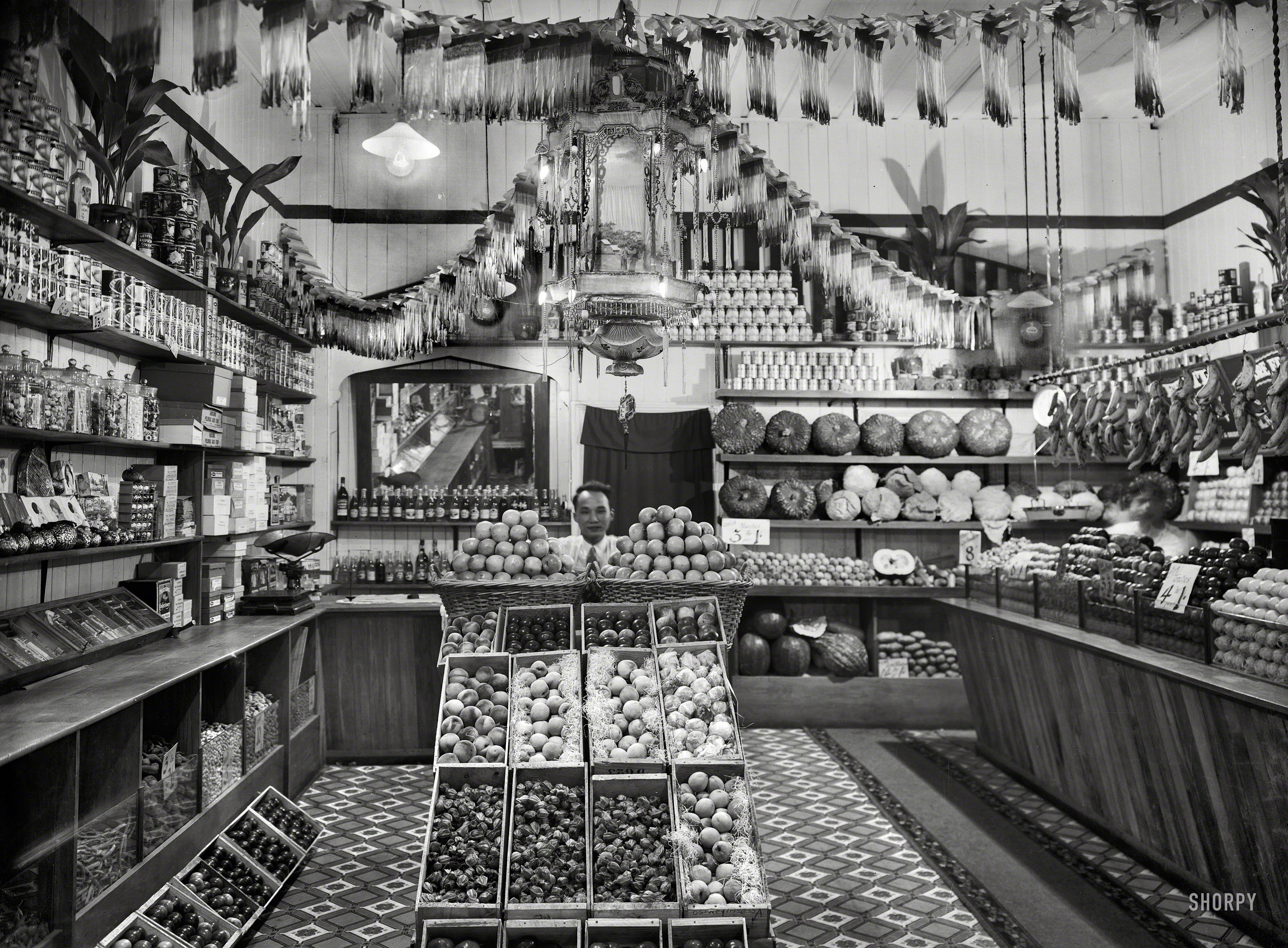 Circa 1920s New Zealand. "Greengrocery, probably Taranaki region. Chinese shopkeeper with baskets of apples and boxes of peaches, cape gooseberries and other fruits." Glass negative by John Reginald Wall. View full size.