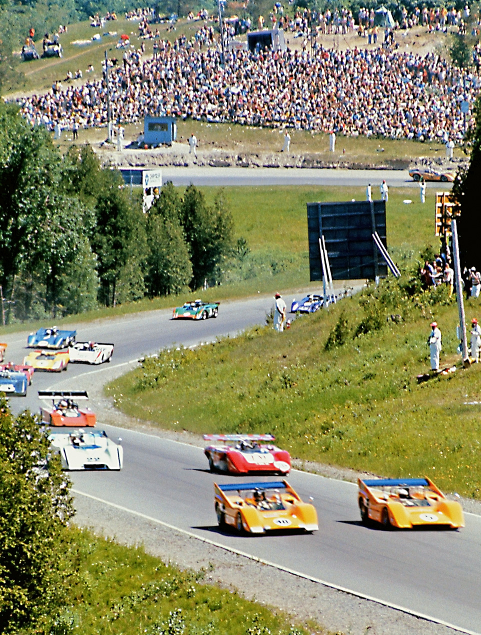 The pace lap of the 1970 Watkins Glen CanAm race, with two McClaren Chevys leading the field. Lots of fans enjoying a sunny, hot race day. View full size.