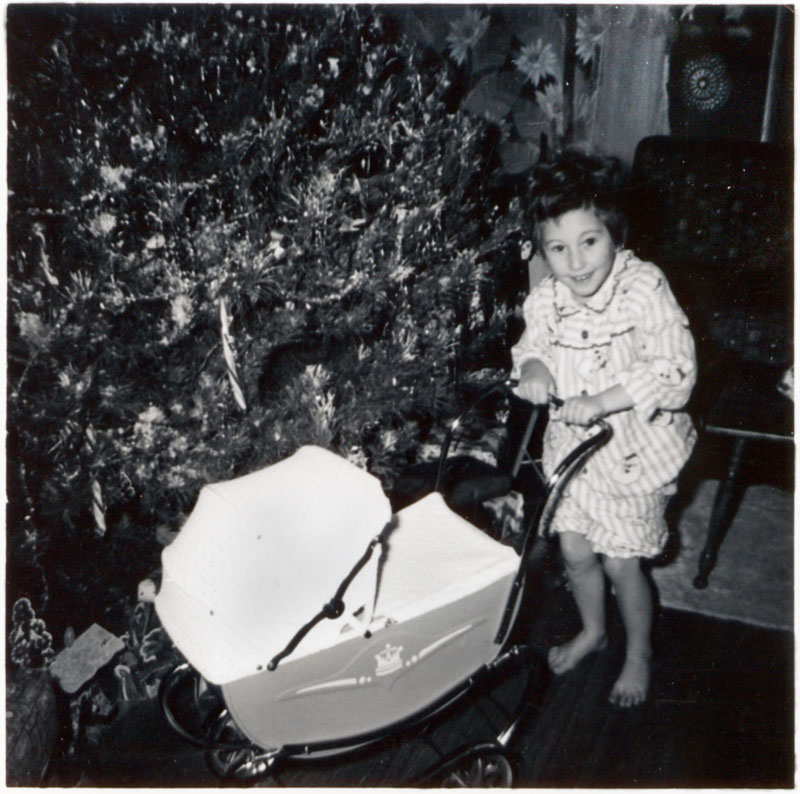 Me (age 5 in my Santa jammies), Christmas morning, Kemptville, Ontario. I noticed the paper cut-out Nativity scene under the tree which I am about to take out with my new pram.
I still remember the tree. It was so wide the boy (one of the students from the high school where my mother taught) who brought it into the house could barely get it in the door. It was also suffering from a tree blight which had affected all the trees in the area. My mother started a trend in the village that year when she painted the brown tips of the branches with silver spray-paint. View full size.
