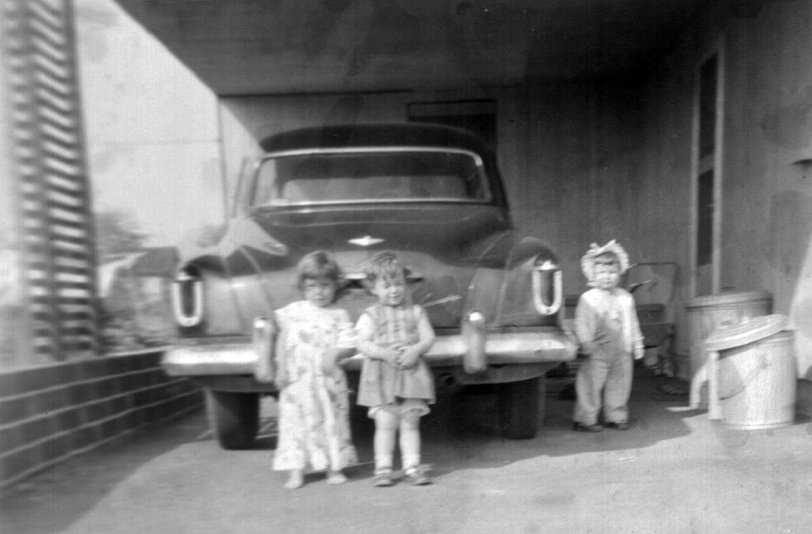 My first car was a 1952 Studebaker Commander because that is the car my parents drove me home from the hospital in after I was born in 1954. It was the only car my family had until November of 1959. It was a four door sedan with the new V8 engine (that had debuted in the 1951 bullet nose version of this car) and a heater. Every other option available for this car was declined: no radio, carpets, wheel covers, decorative trim. It had only a manual transmission. Plus it never had a garage. This Levittown, Pennsylvania carport was as close as it came. By the time my father sold it to some neighborhood teenager in 1962 it had chrome pits the size of cat teeth.
Here you see me (center child) and two playmates, our “gorgeous” zinc plated trash cans, kitchen door, my baby stroller, and my big, black car.
