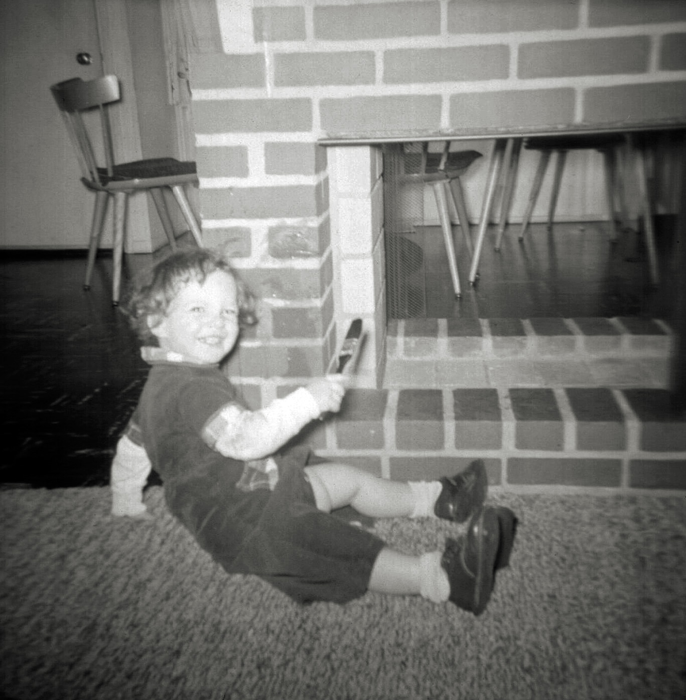One of my absolute favorite childhood games was painting the fireplace bricks with water. If you don’t believe it is a good way to entertain a pre-school child, give that child a paint brush, (such as the one in my hand) and a cup of water, and let them loose on any porous surface that changes color, such as a cement patio, driveway, stones, or bricks. You can barely see the areas I painted, to the right of my face, but you can certainly see my glee.

That unique-to-Levittown crawl-through fireplace which connects the living room (where I am sitting) to the kitchen is also in view. Beyond it are the mid-century modern Paul McCobb kitchen chairs and table my parents bought in 1953 to furnish their then-brand new house. Photo was taken with an Argus 75 box camera and sure-to-wash-out-all-faces flash bulb by one of my parents. Scan was made from a negative. View full size.